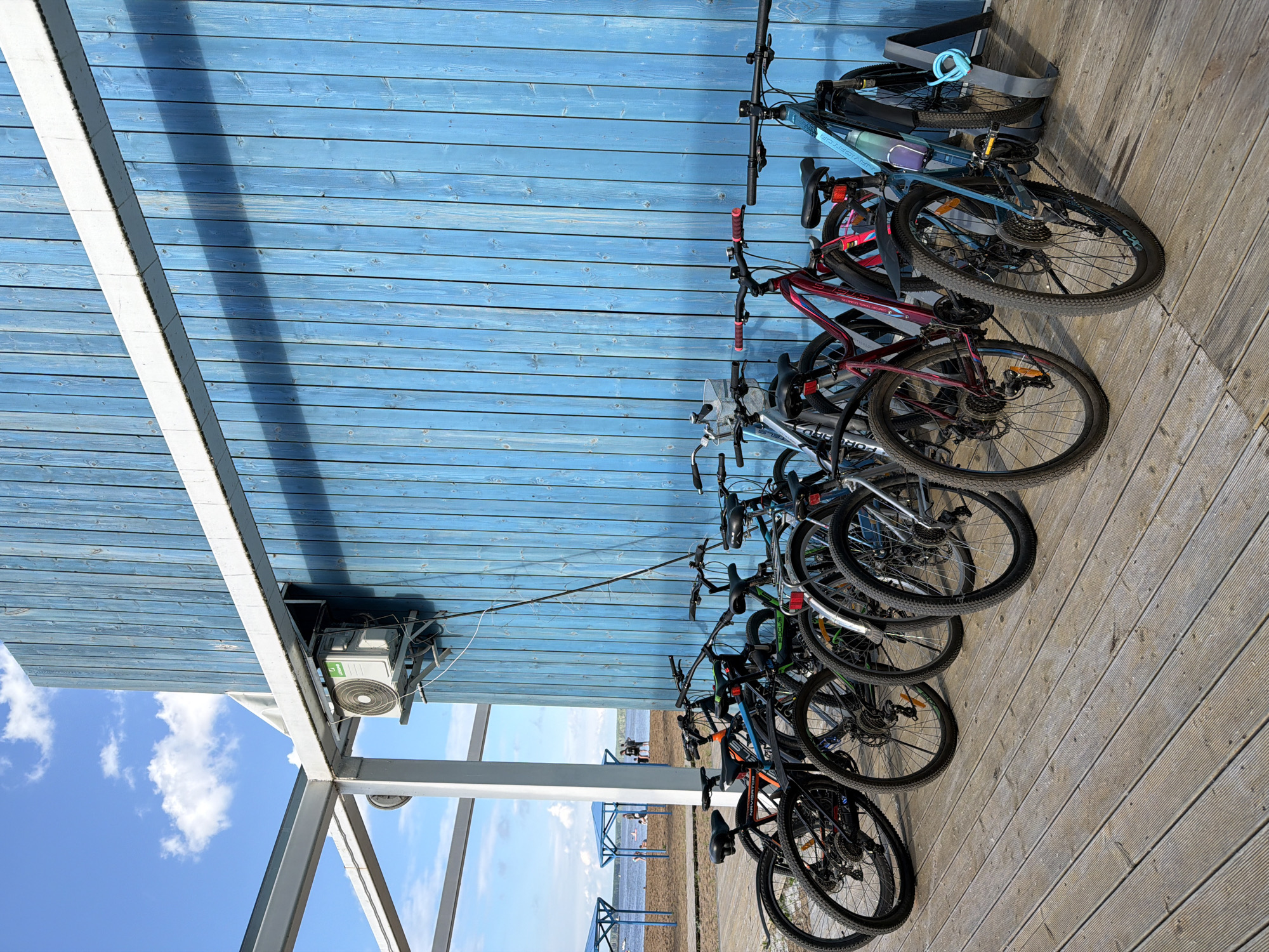 This is a medium shot, eye-level, outdoor photograph taken on a sunny day in Chaikovsky, Russia. In the foreground, a row of bicycles is parked on a wooden deck. The bicycles are of various colors, including black, red, green, and blue, and some have racks and water bottles attached. The wooden deck is made of light-colored planks laid horizontally. In the background, a bright blue wooden wall stands vertically, with a white horizontal beam running across its top. An outdoor air conditioning unit is mounted on the wall. Beyond the wall, a beach with sand and people can be seen, with a body of water and a hazy sky with scattered clouds in the distance.