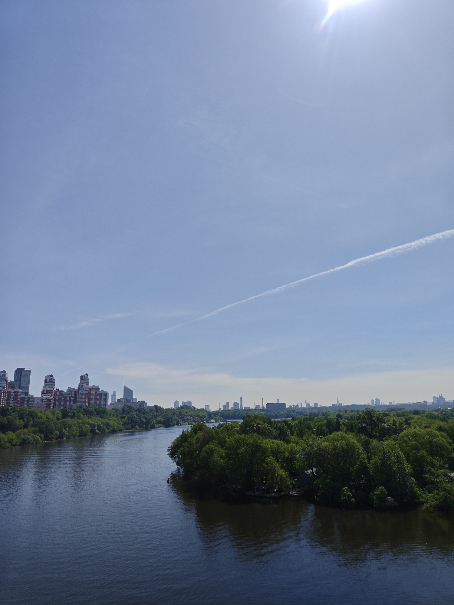 A wide-angle shot reveals a river flowing between banks lined with lush green trees and foliage. On the left bank, there is a cityscape with buildings and skyscrapers. In the distance, more buildings are visible on the horizon. The river appears calm and reflective. The sky is light blue and mostly clear, with a single contrail stretching across the upper part of the frame. The sun is visible as a bright light at the top of the frame. The location is the city of Khimki, Russia.
