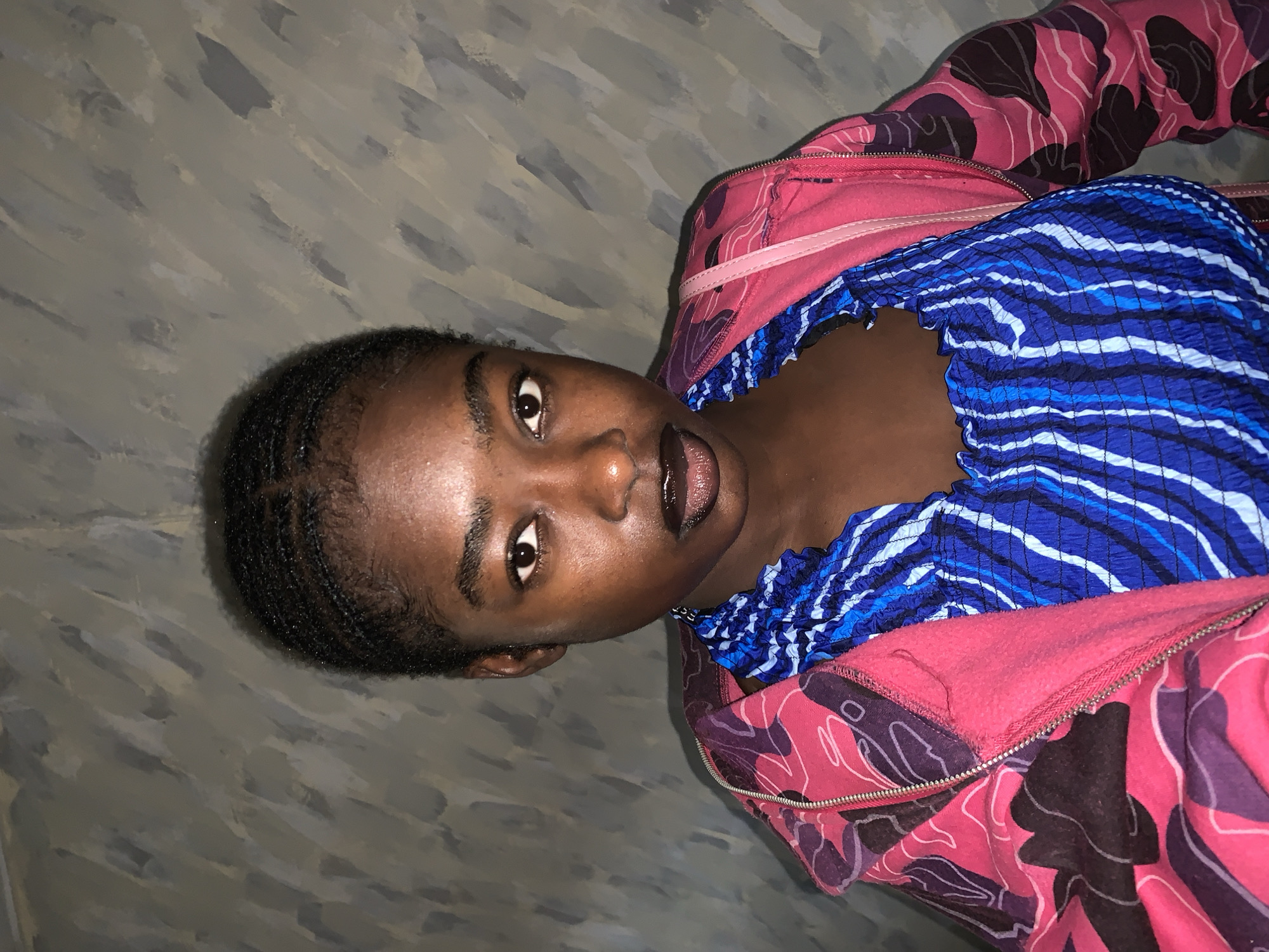 A young Black woman is depicted in a close-up, high-angle shot. She is wearing a blue and white striped top and a pink and purple patterned zip-up hoodie. Her hair is short and dark. The background is a textured wall, possibly painted. The image was taken in Rafin Par, Nigeria.