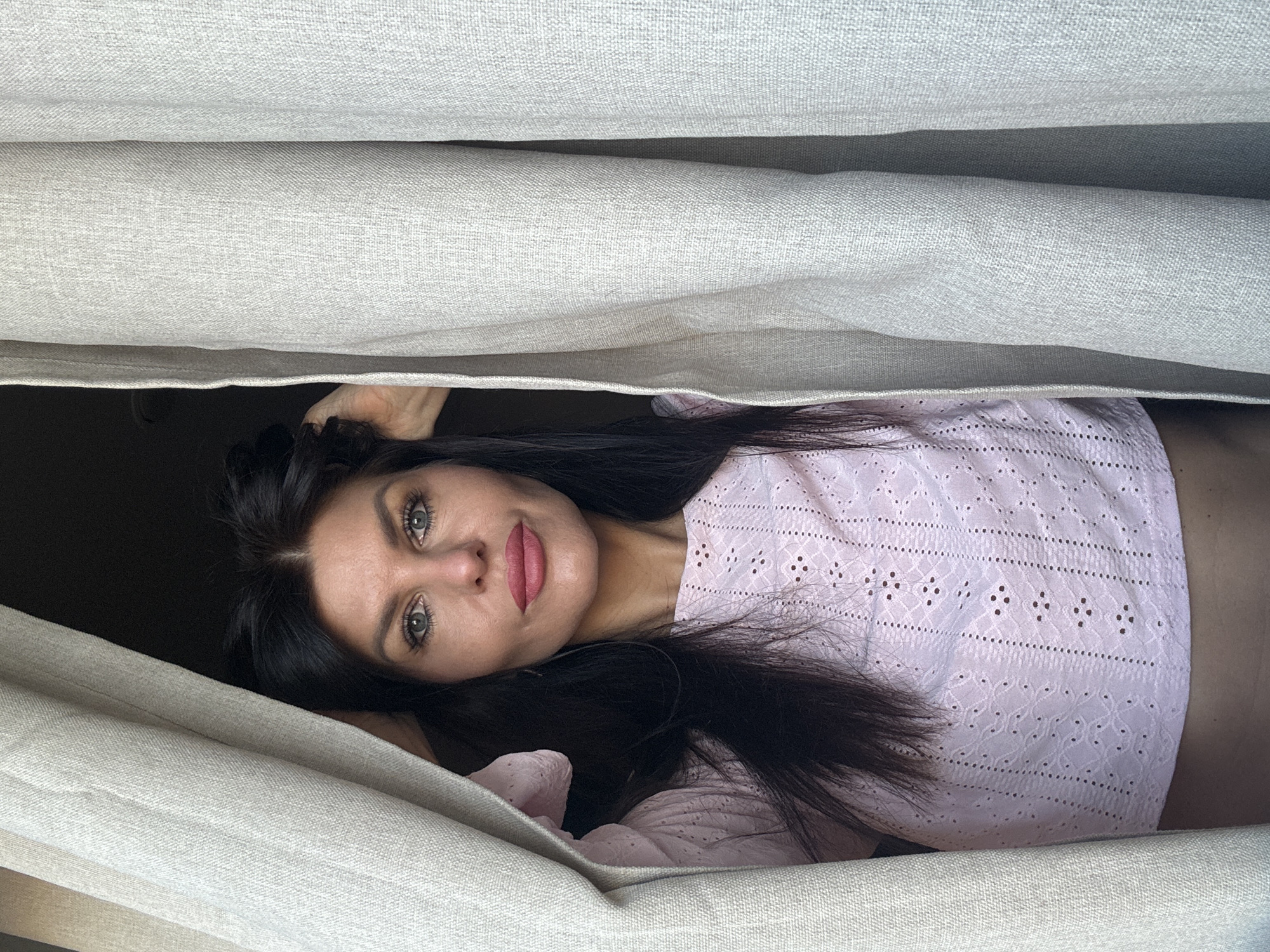 A woman with long, dark hair and light eyes is pictured lying down, peeking out from between two textured, light beige fabric panels, likely curtains or thick blinds. Her head is positioned to the left in the frame, and she looks directly at the viewer with a calm, direct expression. She is wearing pink lipstick and a light pink top featuring intricate white eyelet embroidery. Her dark hair is spread around her head. A dark, indistinct background is visible behind her. The fabric panels create a narrow, horizontal opening through which she is visible, suggesting she is either hidden or playfully revealing herself. The lighting is soft and even, without harsh shadows. No text is visible. The location context is Chaikovsky, Russia.