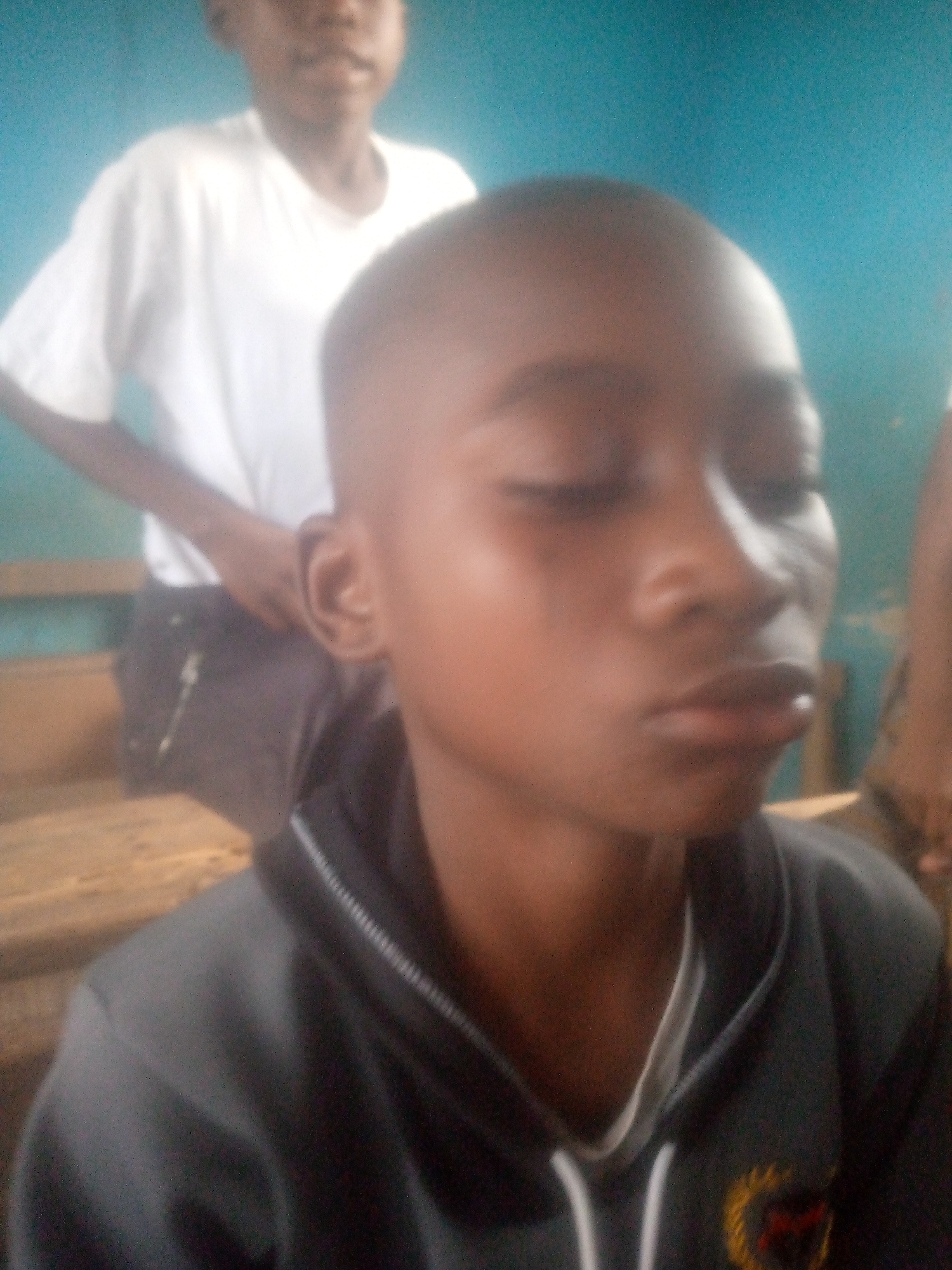The image depicts a close-up of a young, dark-skinned boy with close-cropped hair and closed eyes. He is wearing a dark grey hoodie with a white collar visible underneath. The setting seems to be a classroom. Behind him, a second young boy wearing a white T-shirt stands on what appears to be a wooden bench. The backdrop is a pale blue wall. Given the prompt, the location is presumed to be in Ikorodu, Nigeria.
