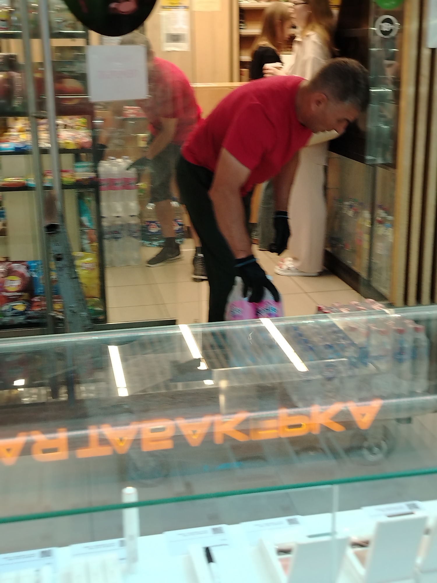The image captures a scene inside a store, likely a supermarket or convenience store, based on the shelving units stocked with goods and the presence of a refrigerated display case in the foreground. In the mid-ground, a man wearing a red t-shirt and dark gloves is bending down, holding a pack of pink-wrapped items, possibly drinks or snacks. He appears to be reaching into or placing items into a display. Behind him, two women are visible, one in a light-colored garment and another in a dark top. The background shows more shelving with various products. The foreground features a glass display case with several rows of bottles, some appearing to be water bottles. The lighting suggests an indoor environment with overhead lights. The reflection on the glass display case in the foreground shows large, orange, block letters that read "HABATRAKEBY". The scene is static, with no discernible movement captured. The floor appears to be tiled.