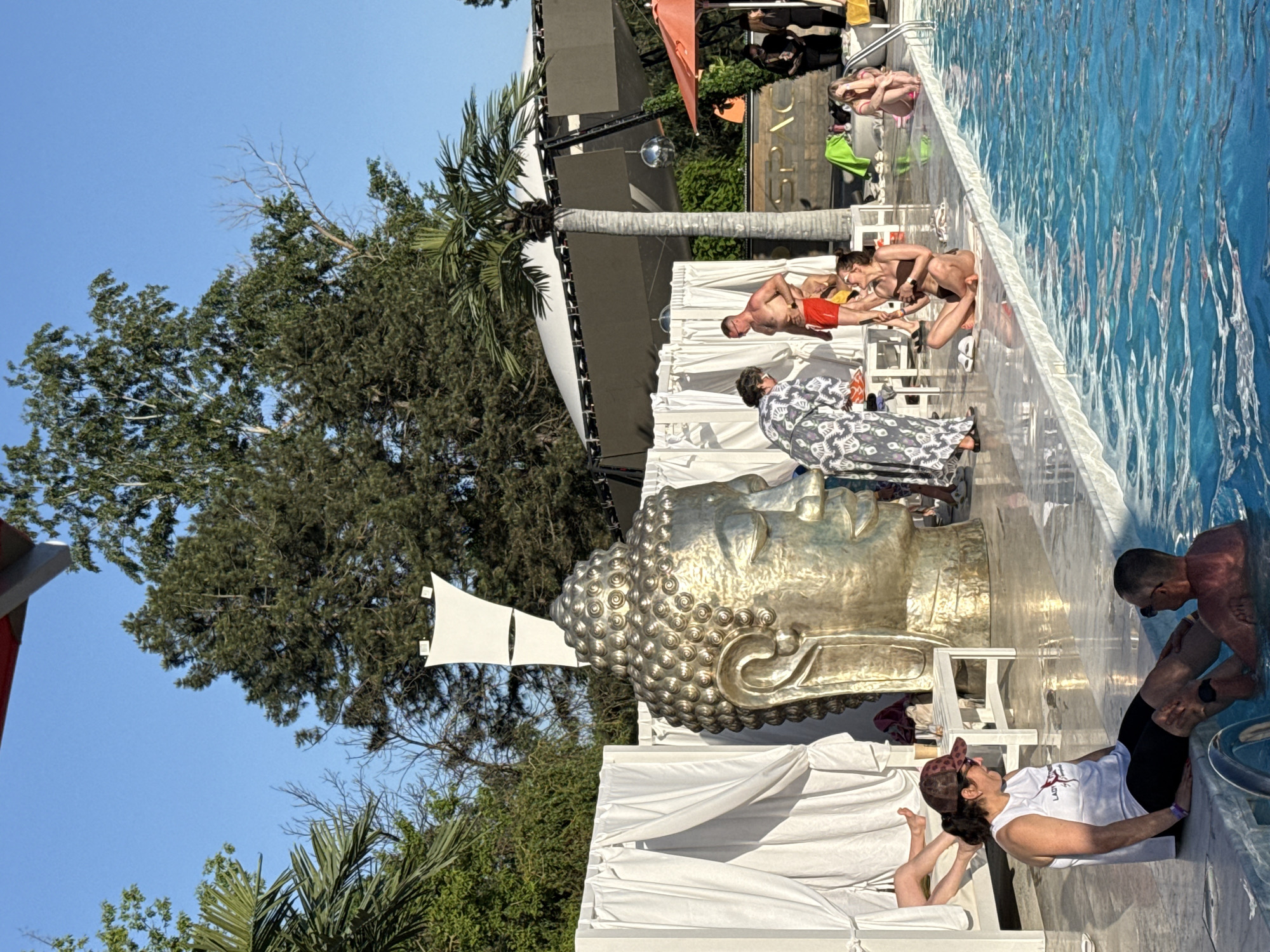 The image depicts a poolside scene in Rostov-on-Don, Russia. A large, golden Buddha head sculpture is positioned near the pool. People are sunbathing on lounge chairs, relaxing by the pool, and swimming. A stage is visible in the background, with audio equipment and a disco ball. Vegetation, including trees and palm trees, surrounds the area. The sky is clear and blue.