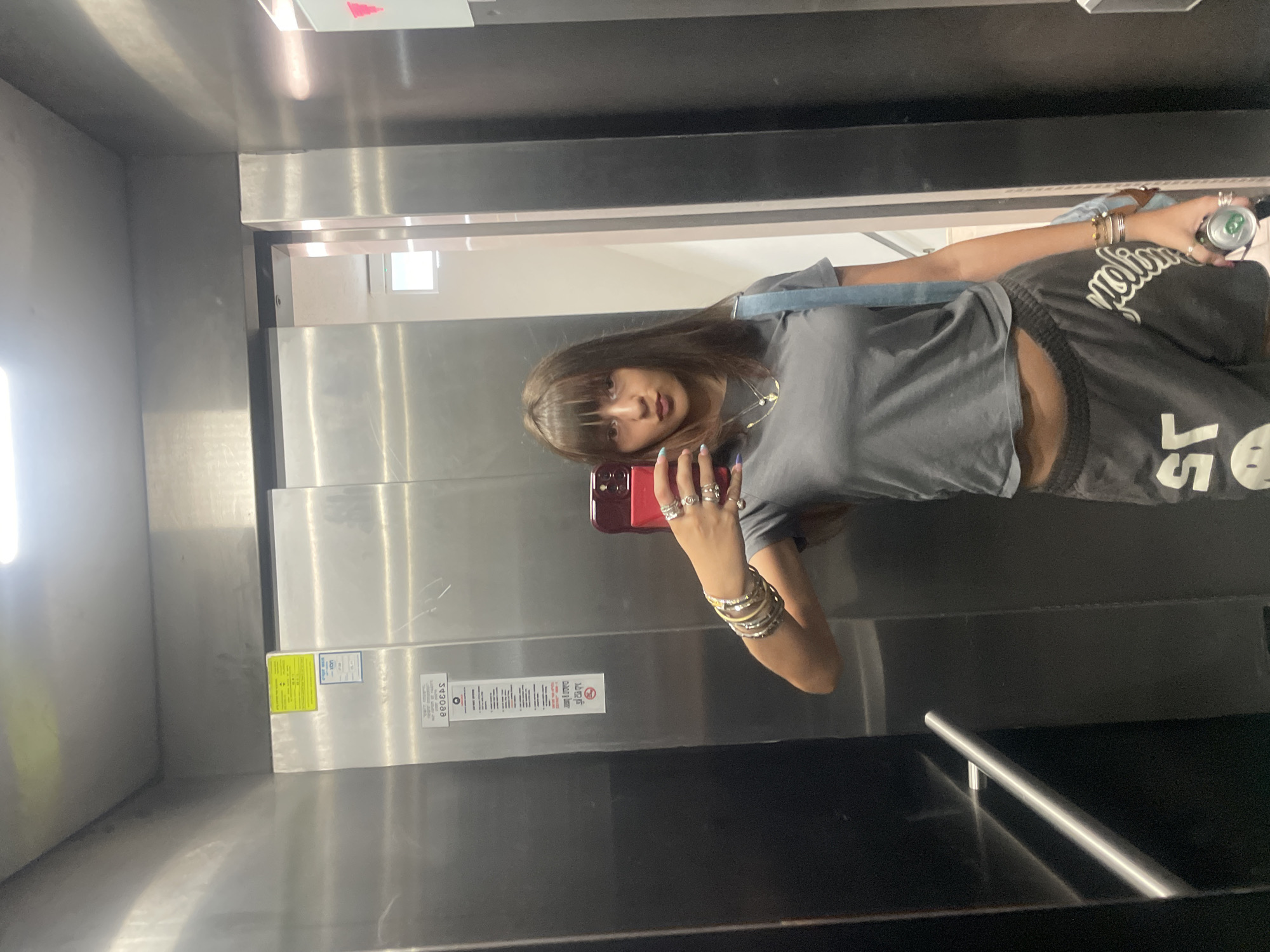 An individual stands in an elevator, taking a selfie with a red-cased smartphone. The person has brown hair with bangs, wears a grey cropped t-shirt, and dark grey shorts with white lettering. They are adorned with multiple bracelets on their left wrist and several rings on their fingers. A light blue strap is visible across their right shoulder, and they are holding a green and silver can in their right hand. The elevator features stainless steel walls and a metal handrail on the left side. A sign with text in Hebrew and English is affixed to the wall. The floor outside the elevator is visible through the open doors, showing a brightly lit hallway with a window. The image appears to have been taken in Raanana, Israel.