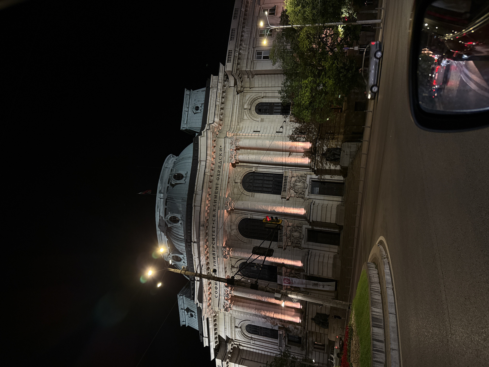 The image captures the architectural facade of Sofia University "St. Kliment Ohridski" in Sofia, Bulgaria, at night. The building's stone exterior is illuminated with warm lights, accentuating its columns, arches, and dome. A street runs alongside the building with a roundabout. There is a car parked on the road and a traffic light is visible on the right of the facade. In the top right corner of the image, a car side mirror reflects the traffic.