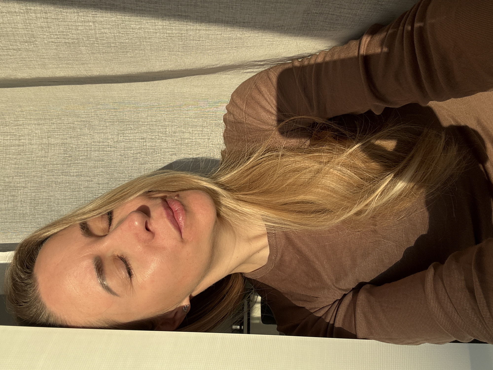 A blonde-haired woman is lying down. The scene appears to be indoors, possibly in the city of Chaikovsky, Russia, though there is no definitive evidence of this. She is wearing a brown, long-sleeved shirt. The environment includes a gray fabric surface in the background and a white, flat surface on which the woman's head rests.