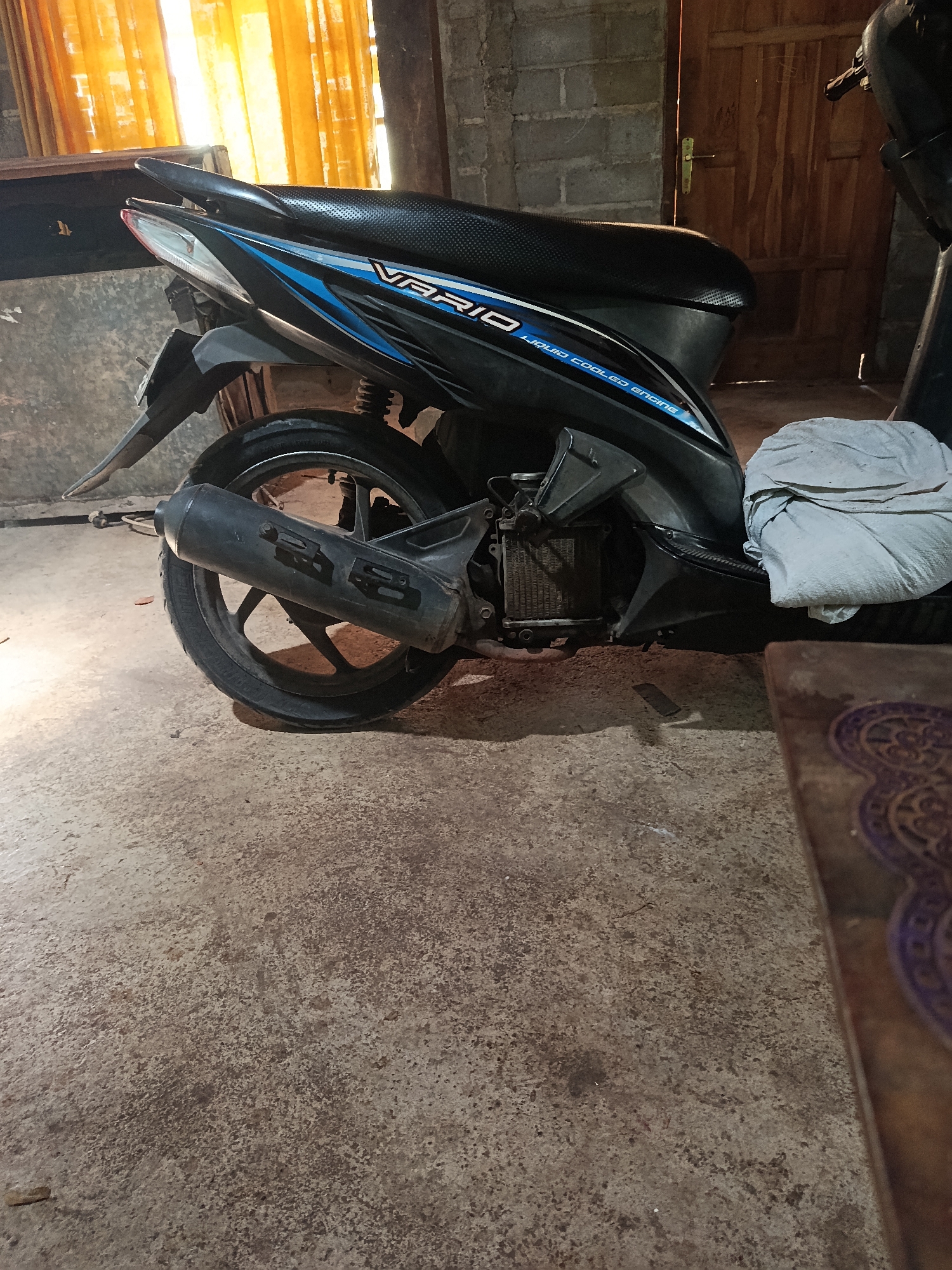 An indoor scene in Karangmojo, Indonesia features the rear section of a black and blue scooter parked on a concrete floor. The scooter has a textured black seat and a blue and white decal that reads "VARIO LIQUID COOLED ENGINE". A grey cloth is draped over the rear of the scooter. To the left of the scooter, sheer orange curtains are visible through a window, letting in sunlight. The walls appear to be constructed from cinder blocks and wood. A wooden door with a brass handle is partially visible on the right side of the image. A portion of a wooden table with a decorative pattern is in the foreground.