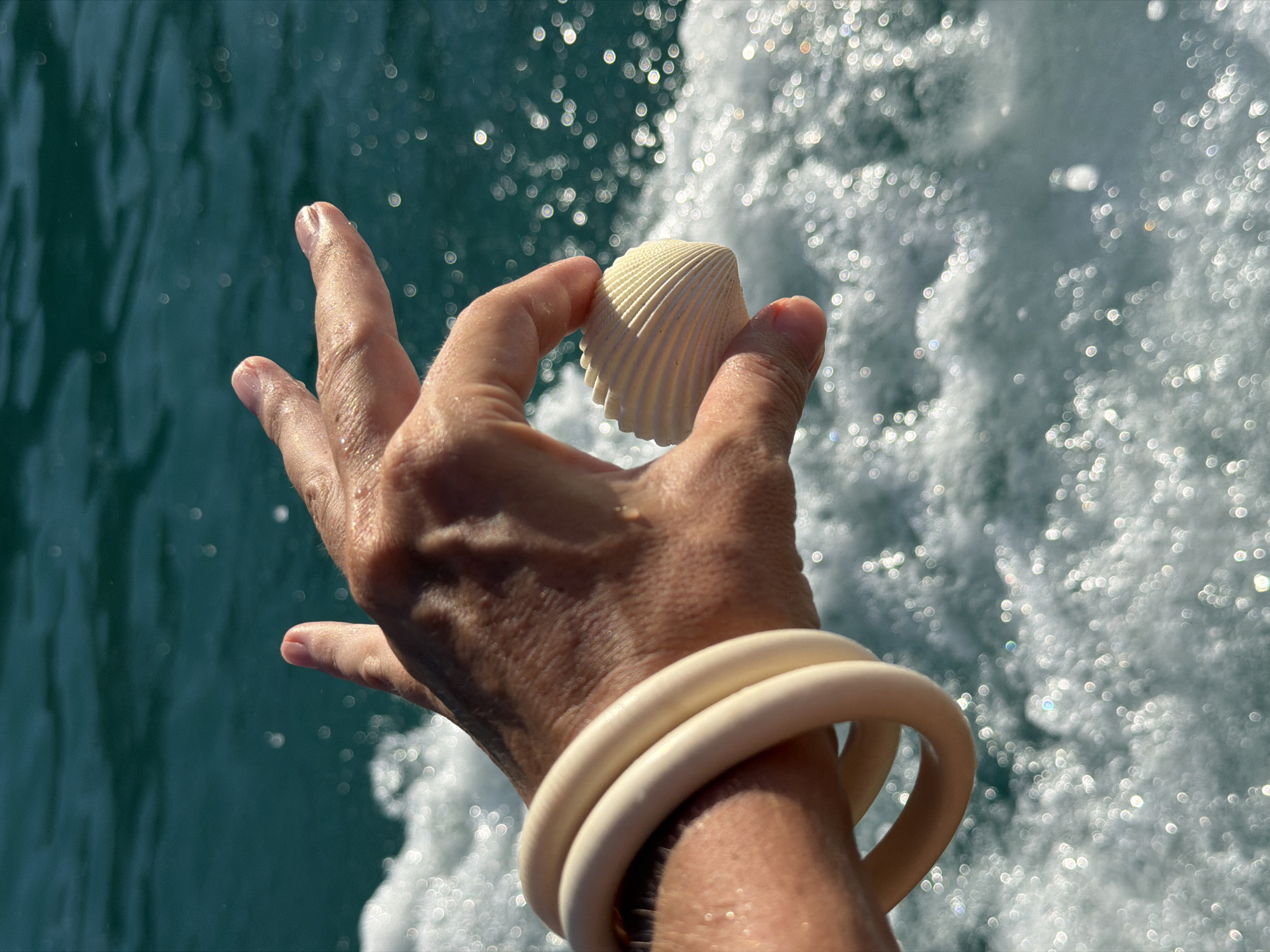 A person's hand, adorned with two cream-colored bangles, holds a small, ribbed seashell against a backdrop of sparkling ocean water. The hand is positioned centrally in the frame, with fingers slightly curved to cradle the shell. The sunlight glints off the water, creating numerous bright, circular highlights that create a bokeh effect. The water itself is a deep turquoise, suggesting it is either the ocean or a large body of water.
The activity appears to be a moment of leisure, possibly on a boat trip, as indicated by the movement of the water and the spray. The bright sunlight and the sparkling water suggest a clear, sunny day, likely during daylight hours. The presence of the seashell and the ocean setting imply a tropical or coastal location. The image conveys a sense of peace and enjoyment of nature. There is no visible text in the image. The location context provided, Baan Thong Krut, Thailand, aligns with these visual cues of a sunny, tropical beach or coastal environment.