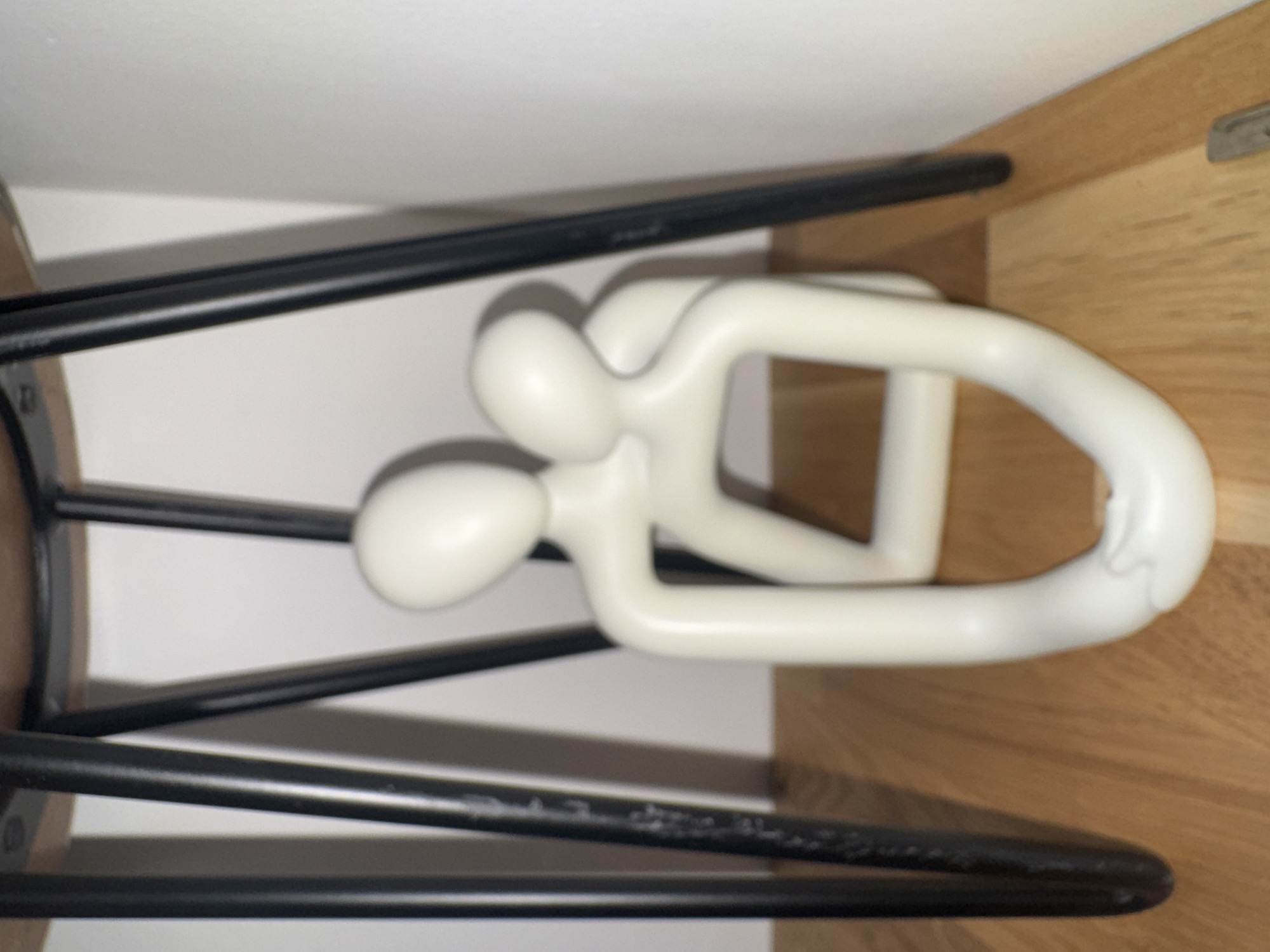 The image shows a white abstract sculpture of two figures in an embrace, placed on a wooden floor beneath a table with thin black legs. The sculpture has a smooth, matte finish and depicts intertwined humanoid forms. The background consists of a plain white wall and the underside of the table, which appears to be made of wood with metal legs. The scene is indoors, suggesting a domestic or indoor public space. There is no information in the image to indicate a specific location in Dacorum, United Kingdom.