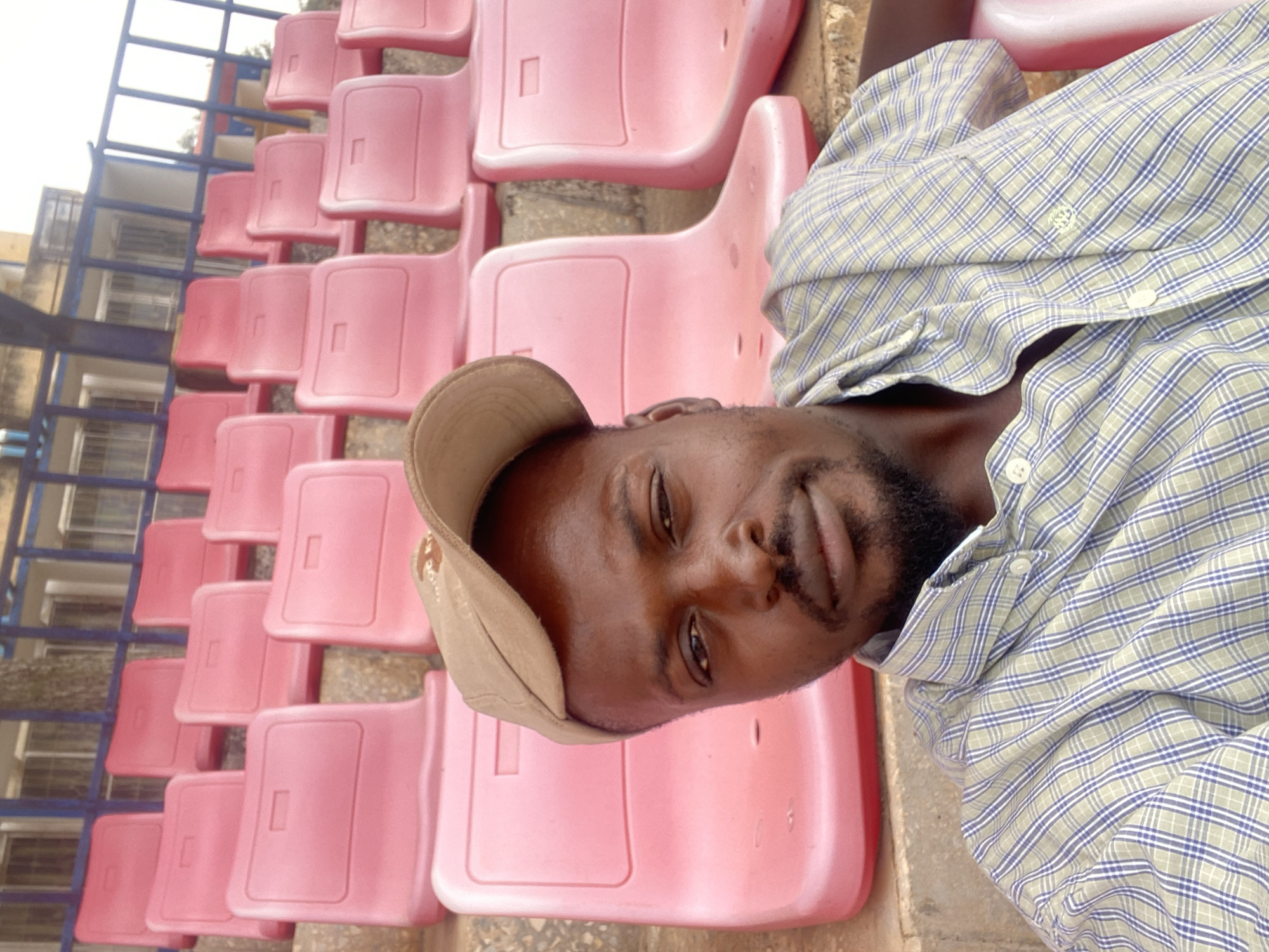 This image, taken in Bukuru, Nigeria, features a man posing for a photograph in an outdoor stadium or arena setting. The man, seen in a close-up, is wearing a light-colored, long-sleeved plaid shirt with a pattern of light green/beige, blue, and white lines. He also has a tan, soft-brimmed cap on his head and a beard. He looks directly at the camera with a neutral to slightly contemplative expression.
Behind the man, numerous rows of empty, bright pink plastic stadium seats extend into the background, defining the location as a public venue. Beyond the seats, blue metal railings or support structures are visible, and further back, a building with windows can be partially seen. The lighting appears to be natural daylight, without strong shadows, suggesting either an overcast day or softer light conditions like early morning or late afternoon. No specific activity is captured other than the man posing, and there is no visible text in the image.