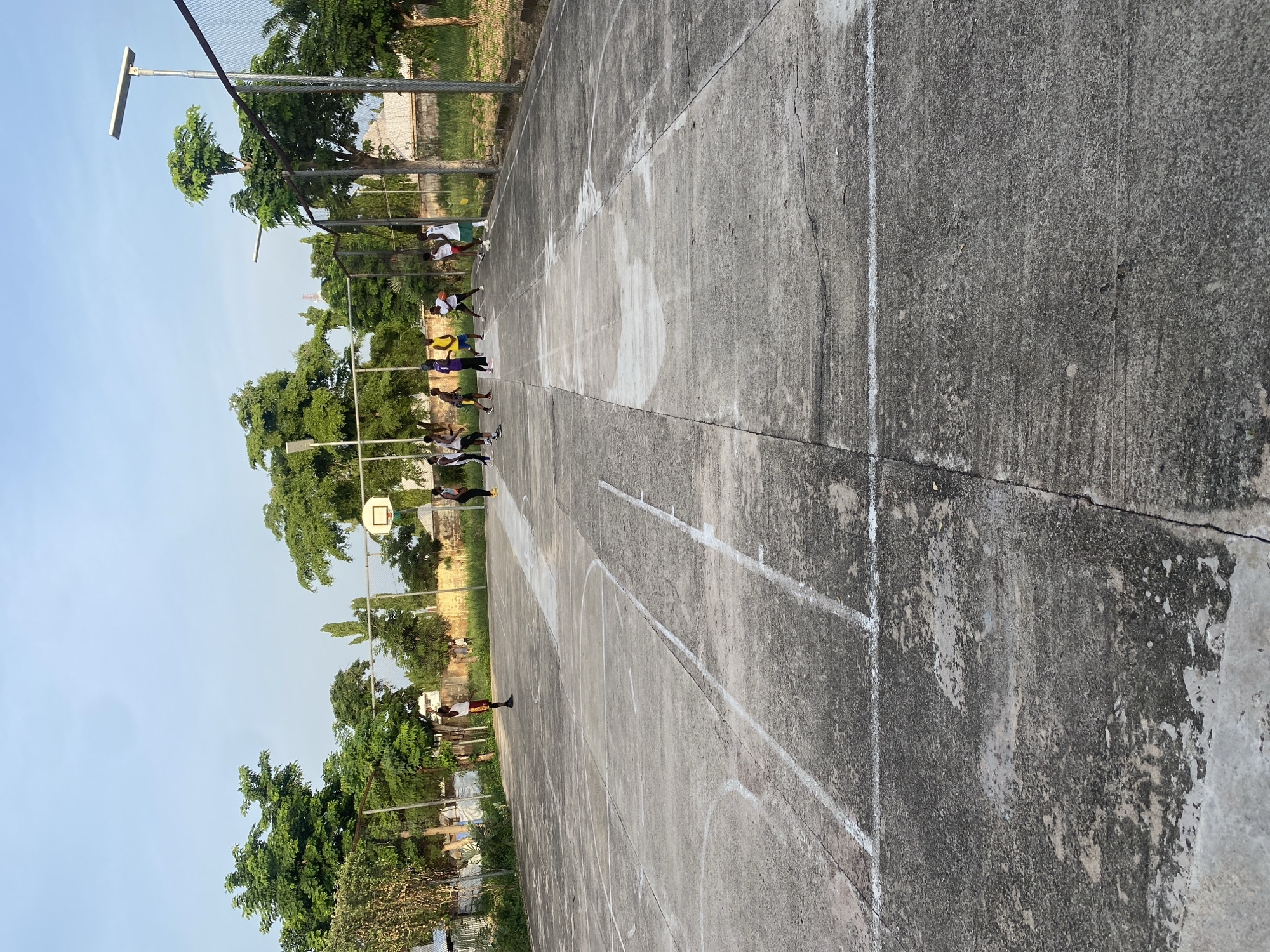 An outdoor basketball court in Jalingo, Nigeria is depicted in the image, with a group of young people playing a game. The court is made of concrete and has visible white lines marking the boundaries. A basketball hoop with a net is visible in the background. The surrounding environment includes trees, a fence, and what appears to be a solar-powered street light. The sky is clear and blue. The people are dressed in athletic wear and are actively engaged in the game, with some dribbling, passing, and shooting the basketball. The overall scene suggests a casual recreational activity taking place in a public space.