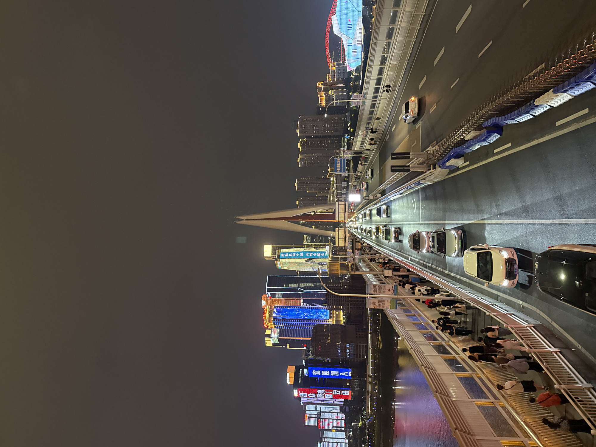 At night, the image shows traffic moving along a bridge, alongside pedestrians. On one side, the bridge has a railing where people are standing, looking out over the scene. On the other side of the bridge, vehicles travel along the roadway. In the distance, tall buildings and structures are illuminated with lights. The image is taken from a high vantage point. The location is Yuzhong District, China.