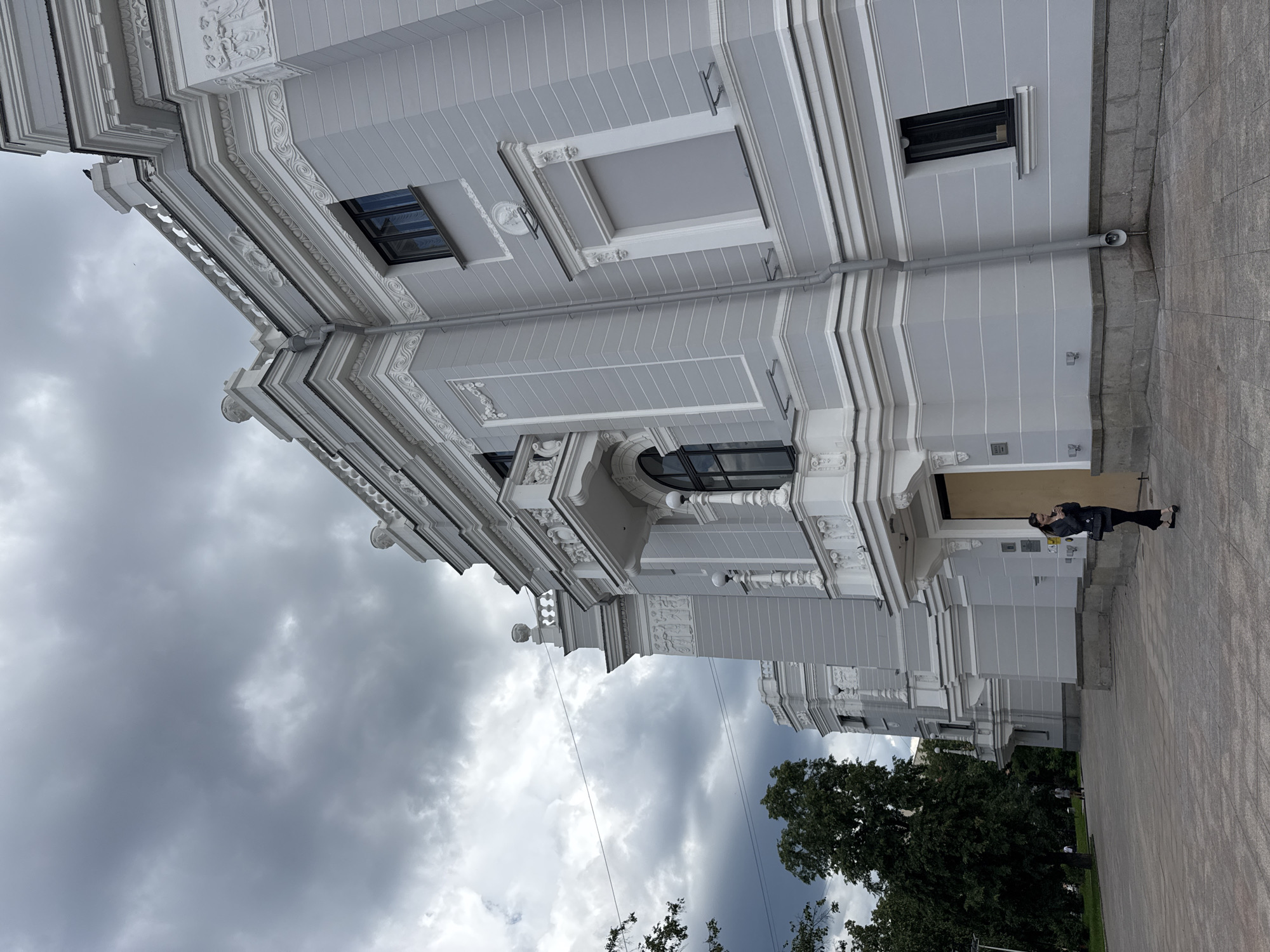 A low-angle, full-shot, outdoor photograph, taken from a paved walkway, depicts a grand, light-gray building with ornate white detailing. The building, located in Yekaterinburg, Russia, has a classical architectural style, featuring columns, balconies with decorative railings, and intricate relief sculptures. Several windows are visible, some dark, others covered with a light gray blind. The sky above is overcast with heavy, dark gray clouds. To the left of the building, several mature trees with dark green foliage are present, and a small portion of a green lawn is visible. Two individuals are in the background on the lawn, appearing to be walking. In the foreground, a lone woman stands on the paved walkway, facing the building. She is wearing dark clothing, including a jacket and pants, and is carrying a black purse over her shoulder. She is holding a small object, possibly a phone, near her face and looking down at it. The lighting suggests it is daytime, possibly before or during rain.