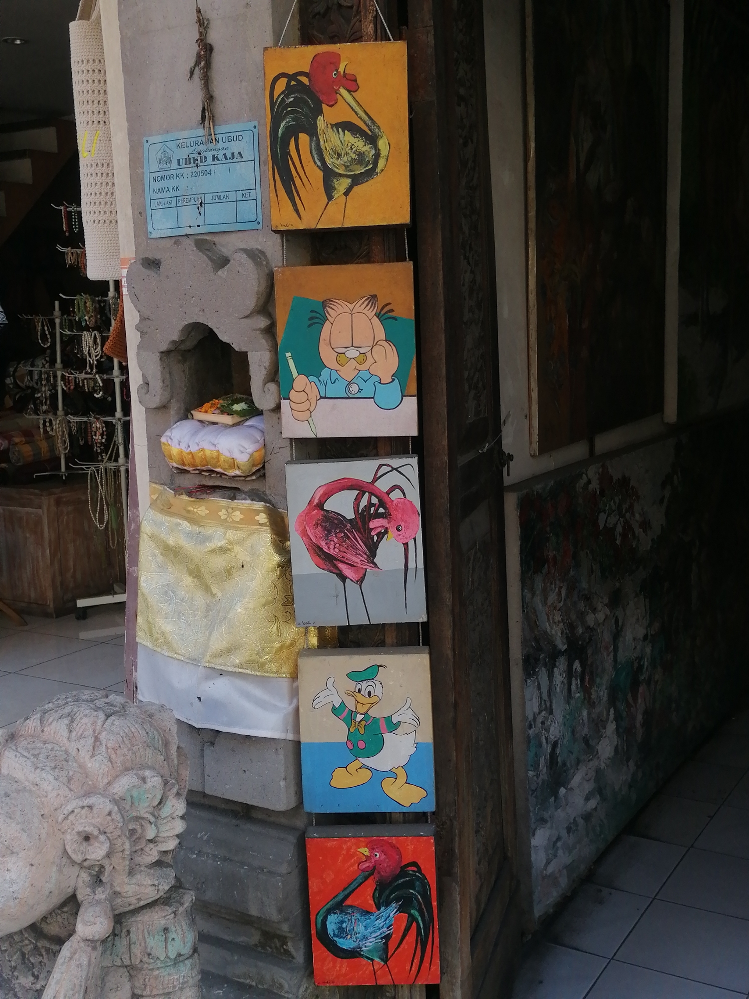 The image shows a collection of paintings displayed on a stone pillar in a narrow outdoor space, likely a shop in Ubud, Indonesia. The paintings depict cartoon characters and roosters, each in a small square format. They are hung vertically in a row, with a light yellow painting of a rooster at the top, followed by a painting of Garfield, a pink flamingo, Donald Duck, and another rooster in a red and blue palette at the bottom.
To the left of the paintings, there's a woven bag hanging and a display of various necklaces and trinkets on racks. A stone sculpture of a mythical creature's head is visible in the lower left corner. The pillar itself is decorated with a white cloth draped with a gold patterned fabric. Above the paintings, a blue sign with Indonesian text is affixed to the pillar. The background on the right reveals a dark, textured wall and a tiled floor. The overall impression is that of a roadside stall or shop selling local art and crafts.