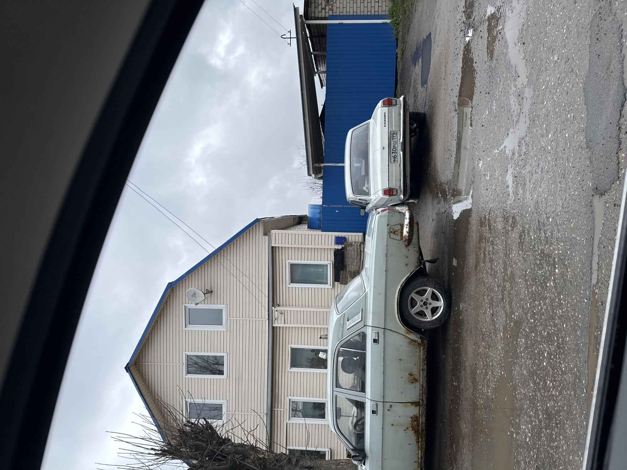 Observed from within a vehicle, the scene depicts a residential area in Ново-Никольское, Russia. Two cars, a light blue sedan with rust and a white sedan with license plate "M630PO 199", are parked on a wet, asphalt road. Adjacent to the road is a two-story, light-colored house with blue trim. To the right of the house, a blue metal building with a gray roof is visible. The sky is overcast, and tree branches are visible in the lower left.
