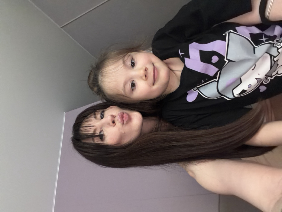 An adult female and a young girl are depicted in a close-up indoor shot, originating from the city of Perm, Russia. The adult female, positioned on the left, has long, straight dark brown hair with bangs, fair skin, and is making a puckered lip expression towards the camera. She appears to be wearing a light-colored, possibly off-shoulder top. The young girl, positioned on the right and slightly in front, has light brown hair styled in a bun with bangs, fair skin, and is looking at the camera with a subtle, closed-mouth smile. She is wearing a black long-sleeved t-shirt featuring a prominent graphic design in light purple and grey, depicting an animated cat-like character surrounded by hearts and geometric shapes. A black hair tie is visible on her left wrist. The background consists of a solid light purple wall on the left, meeting a white ceiling. A different, lighter-colored (grey or white) wall or surface is visible on the right side of the frame, suggesting an interior corner. Both individuals are closely framed, indicating a selfie or intimate portrait.