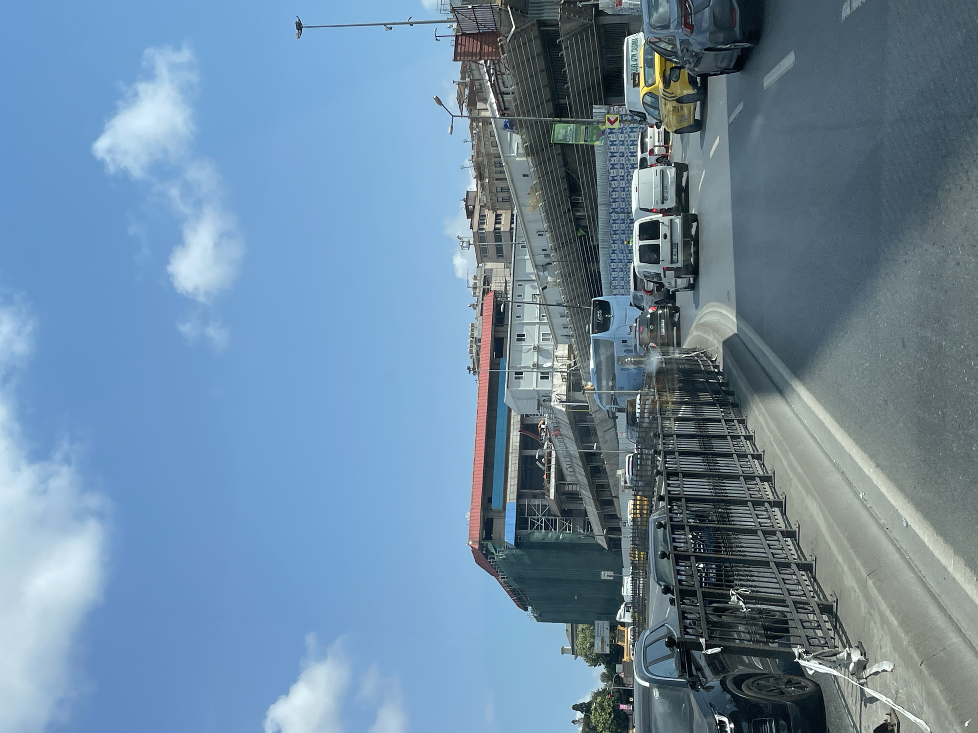The image shows a street scene in Beyoğlu, Turkey, on a sunny day with a blue sky and scattered clouds. The foreground features a multi-lane road with several vehicles, including black and gray cars, white vans, a blue bus, and a yellow taxi. The vehicles are moving in a line, suggesting moderate traffic. A black metal fence runs along the side of the road, separating it from a sidewalk and a construction site.
The construction site in the background is a large building covered in green netting, with scaffolding and exposed structural elements. The building appears to be under renovation or new construction, with a red roof and various levels visible. Other buildings, some residential and some commercial, are visible in the background, with a mix of architectural styles. Streetlights and traffic signals are also present. The overall impression is of a busy urban environment with ongoing development.