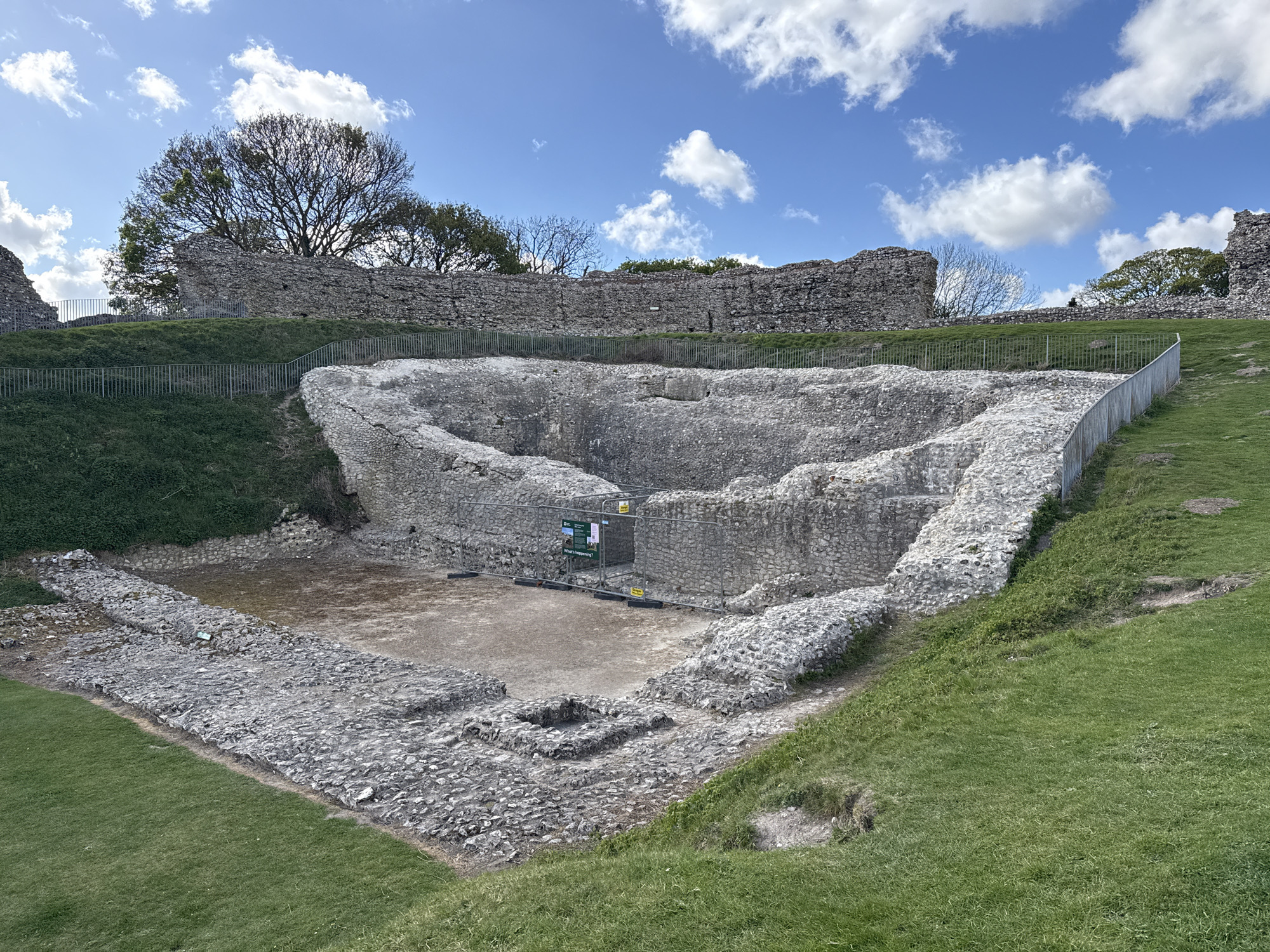 The image captures the extensive stone ruins of a historical structure, likely a castle or fortification, under a clear blue sky dotted with fluffy white clouds. The setting is an outdoor, grassy area, suggesting a historical site or park.
In the foreground and central part of the image, the remains of an excavated or decayed stone structure are visible. This area features low, irregular walls made of light-colored, rough-hewn stones, forming a large, somewhat triangular or semi-circular enclosure. Within this central pit, there are lower, square-shaped stone foundations on a dirt floor. Temporary metal fences, typical of construction or archaeological sites, surround a portion of this central ruin. A small green and white sign is visible behind these fences, partially displaying the text "What's happening?".
Beyond these immediate ruins, higher and more substantial stone walls form a perimeter on an elevated grassy mound. These walls are also constructed from similar irregular stones. Lush green grass covers the slopes leading up to these higher walls and surrounds the entire ruin complex. Several trees, mostly bare-branched with some green foliage, stand behind the distant upper walls, silhouetted against the bright sky. Metal railings, acting as permanent safety barriers, run along the edges of the grassy slopes and around parts of the ruins.
The photograph was taken on a bright, sunny day, likely in the late morning or early afternoon, given the clear light and defined shadows. The overall impression is one of ancient history preserved within a natural landscape, located in the King's Lynn and West Norfolk area of the United Kingdom.