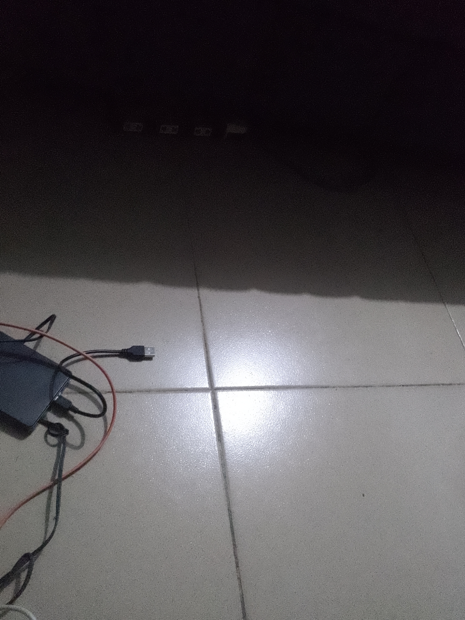 In an indoor setting, a dark-colored electronic device, possibly a power bank or external hard drive, lies on a tiled floor. Connected to the device are several cables, including a black USB cable and a red cable. The floor is illuminated by a light source casting shadows and reflections. The upper portion of the image is dark, with faint outlines of what could be a shelf or furniture. The scene appears to be set in an unknown location within an unknown city.