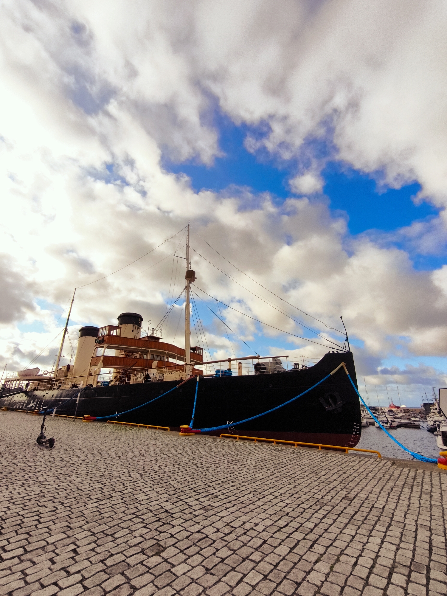 In Tallinn, Estonia, a large black ship named "SUUR HALL" is docked at a harbor. The ship features a multi-level superstructure with wooden accents and two prominent smokestacks. Blue ropes are used to secure the vessel to the dock, which is paved with cobblestones. A black electric scooter is parked on the cobblestone walkway in front of the ship. The sky above is filled with dramatic white clouds against a blue backdrop. In the background, other smaller boats and ships are visible in the harbor.