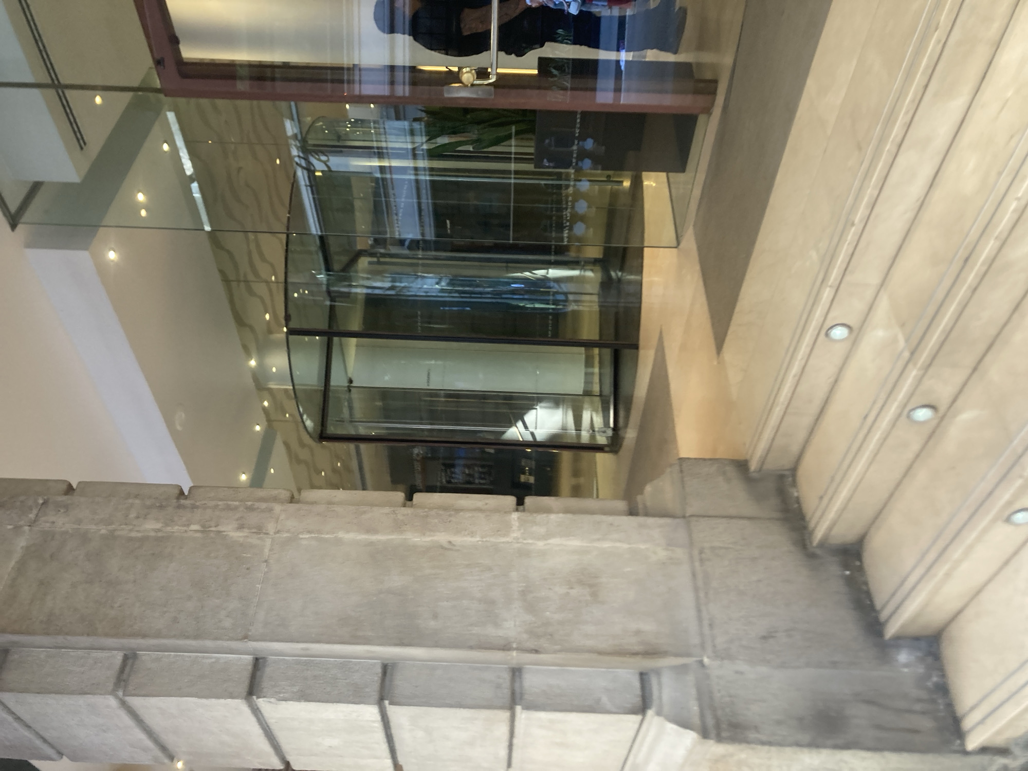 The image depicts the entrance to a building, likely a hotel or a commercial establishment, located in Barcelona, Spain. In the foreground, a wide, light-colored stone staircase leads up to the entrance. Each step is illuminated by a small, circular light embedded in its riser. To the left of the staircase, a large, fluted stone column stands prominently.
The entrance itself features a glass revolving door, through which glimpses of the interior can be seen. Beyond the revolving door, the lobby area appears to be well-lit, with recessed ceiling lights visible. The interior design includes reflective surfaces, likely glass and polished materials, creating a sense of spaciousness. A tall, dark planter with a green plant is positioned to the right of the revolving door, near a glass wall.
To the far right of the frame, a person is visible standing partially obscured by a dark wooden framed glass door, suggesting this is an additional entrance or exit. The person is wearing dark clothing and their silhouette is cast on the glass. The overall environment suggests a modern, upscale building entrance.