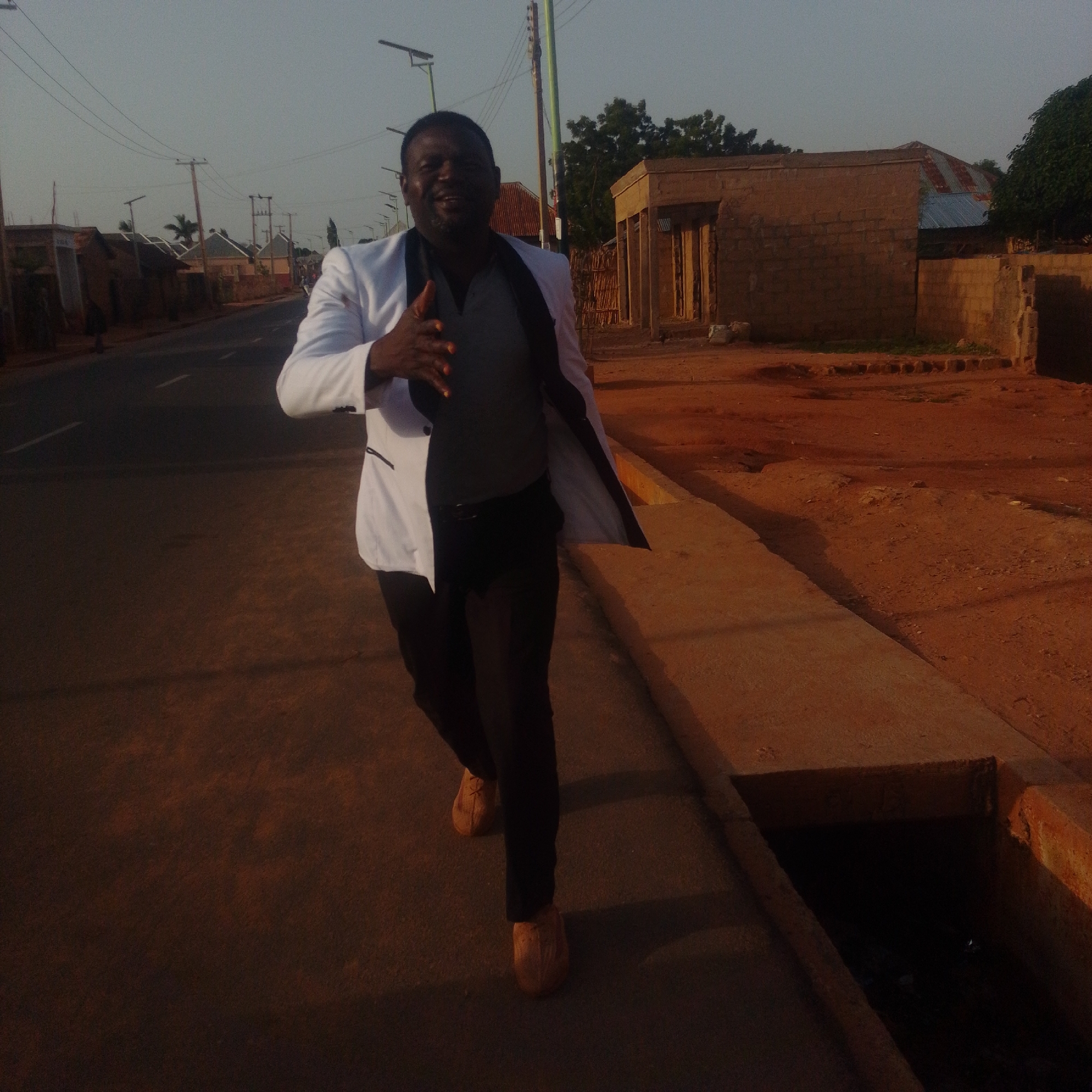 A man in a white blazer, grey shirt, and black pants walks towards the camera on a dirt road next to a paved sidewalk. He is smiling and extending his right hand. In the background, there are buildings, utility poles, and trees under a clear sky. The overall scene appears to be a residential area in a city.