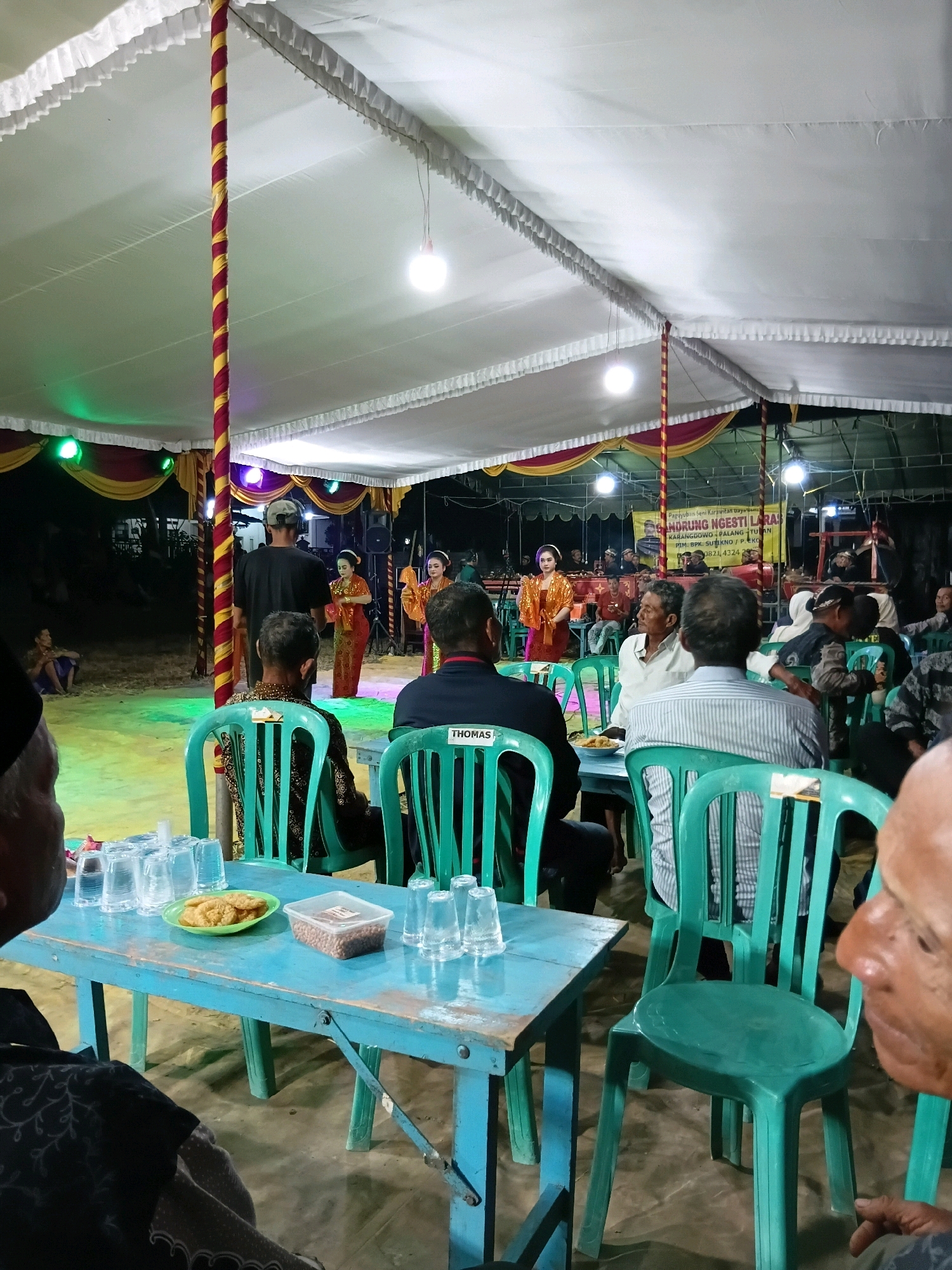 The image depicts an outdoor event, possibly a cultural performance or celebration, taking place under a white tent adorned with red and yellow fabric draping. The scene is illuminated by overhead lights and green and yellow uplighting. In the background, a stage area is set up with several individuals, including two women in traditional orange and red attire, possibly performers. A banner with Indonesian text is visible behind them. In the foreground, a blue table holds several glasses, a plate of food, and a container of peanuts. People are seated on green plastic chairs around the table, facing the stage. Some attendees are in the mid-ground, also seated on chairs. The overall environment appears to be sandy or dirt ground. The event is taking place in Soko, Indonesia.
