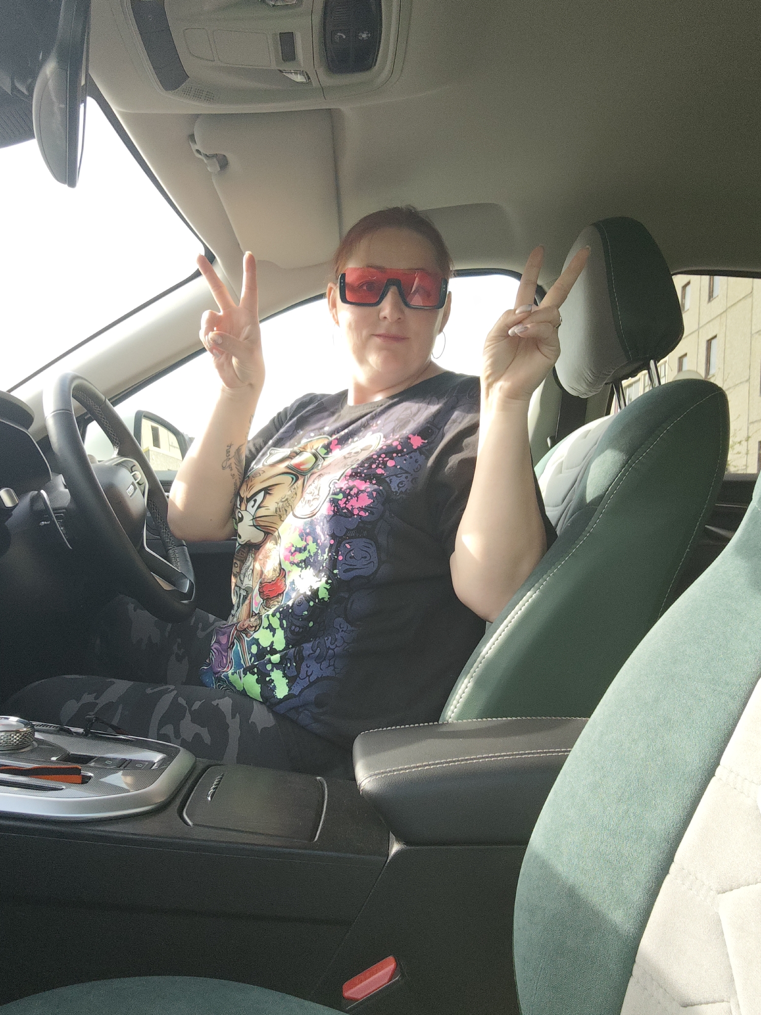 A woman is sitting in the driver's seat of a car, making a peace sign with both hands. She is wearing red sunglasses and a t-shirt with a cartoon character print. The car has green and white suede seats and a modern dashboard. The scene appears to be taking place during daylight hours, as evidenced by the bright sunlight entering through the windshield. In the background, a building is visible, suggesting an urban setting. While it is possible the image was taken in Markovskii, Russia, as indicated in the query, this cannot be definitively confirmed from the image alone.