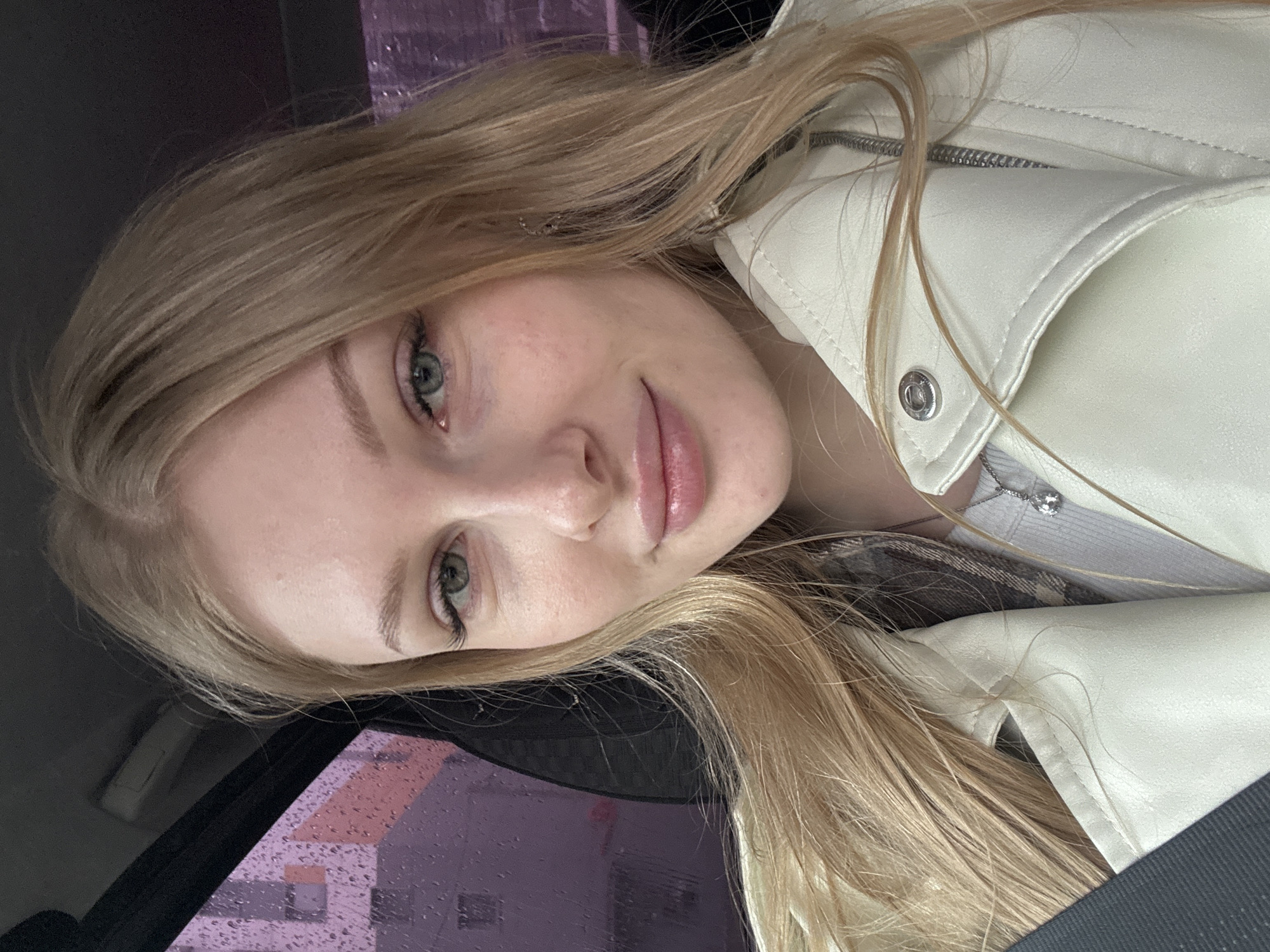 A fair-skinned woman with blonde hair is facing the camera, wearing a white leather jacket and a plaid shirt. She appears to be inside a car, with a rain-streaked window visible in the lower left. Outside the window, there's a blurry view of buildings, possibly in Perm, Russia. The woman has light green eyes and is smiling subtly. A silver necklace is visible beneath the jacket collar. The car interior is predominantly dark, providing contrast to the woman's light skin and clothing.