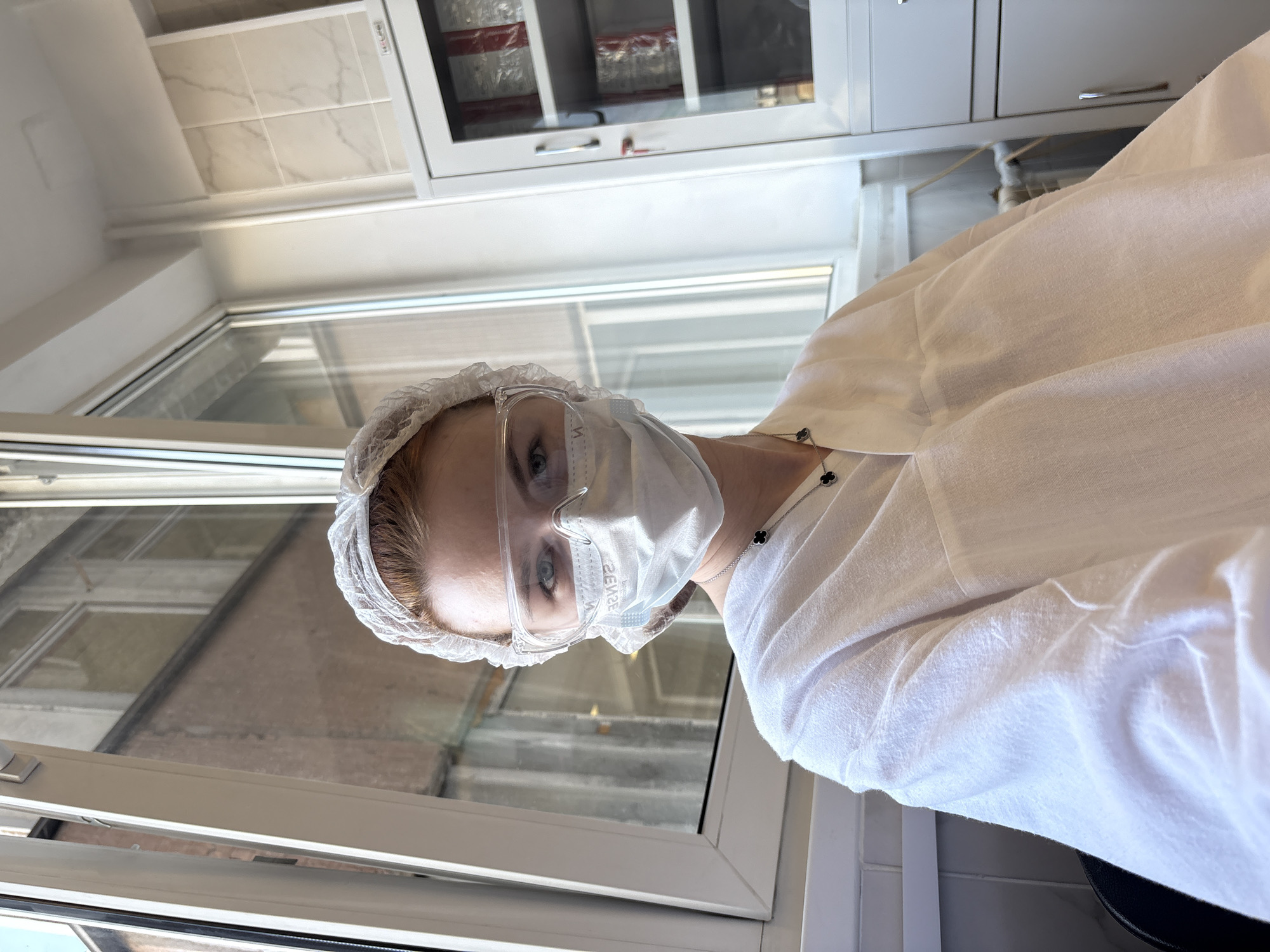 The image captures a person, likely in a professional or clinical setting in Saint Petersburg, Russia, looking directly at the camera. The individual wears extensive protective gear, including a disposable white hairnet, clear safety goggles, and a white surgical face mask that covers their nose and mouth. Their blue eyes are visible and focused. They are dressed in a loose-fitting white smock or lab coat. A silver necklace adorned with black clover-shaped charms is visible around their neck.
The background elements suggest an indoor environment. To the left of the person is a large window with white frames, through which reflections are visible, as well as a glimpse of an exterior building with reddish-brown brick-like textures, typical of urban architecture. Above the person and the window are white cabinets. One cabinet features glass doors, behind which items wrapped in red and clear plastic are stored, possibly medical or laboratory supplies. Above these cabinets, light-colored wall tiles with a subtle pattern, possibly marble, are visible. The scene is brightly lit, indicating it is daytime. On the white surgical mask, faint mirrored text can be observed, which appears to be "SENSE2" when un-mirrored. The overall impression is that of a person ready for work in a sterile or laboratory-like environment.
