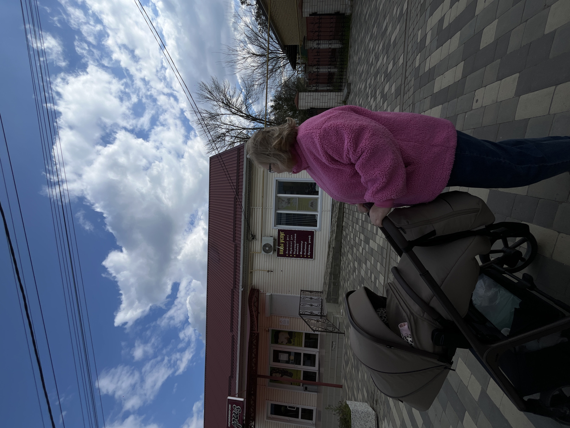 A woman in a pink jacket and blue jeans walks along a brick sidewalk, pushing a beige stroller. To the left is a one-story building with a red roof and beige siding, identified as a barber shop. A sign lists services such as "Ladies' Hall, Men's Hall, Nail Extension, Manicure, Pedicure, Ear Piercing." The shop's name, partially visible, reads "Paris." Overhead electrical wires are visible against a backdrop of cloudy blue sky. The location is identified as Novopokrovskaya, Russia.