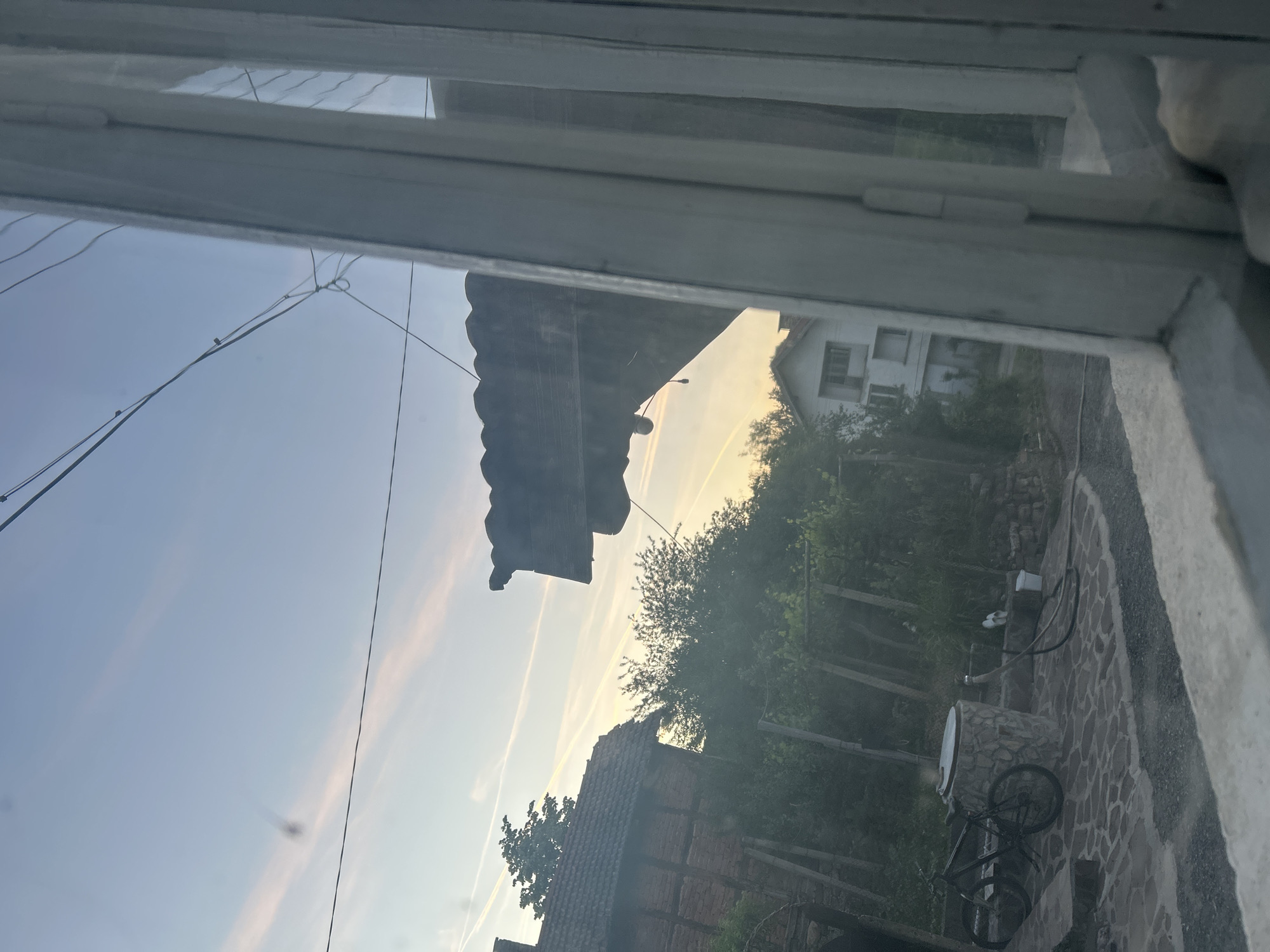 The image is a view through a window, likely in a residential area. Outside, there are several buildings: one with a steeply pitched, tiled roof made of brown bricks, a modern white building with windows, and a structure with a corrugated roof. Trees and other vegetation are visible, and the background sky has a gradient from light blue to yellow. In the foreground, a stone patio and a bicycle are visible. The location seems to be in an unknown city.