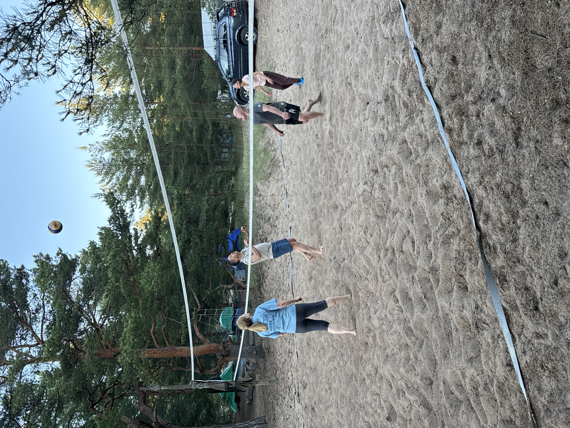 A group of four individuals are playing volleyball on a sandy court in Ozerki (Krasnodolinskaya), Russia. The court is set up with a net, and the players are in various stages of hitting or observing the ball. Two men and two women are present. One woman in a blue t-shirt is positioned on the left side of the net, facing away from the camera. A man in a light-colored polo shirt and blue shorts is on the left side, looking up at the ball. On the right side of the net, a man in a dark t-shirt and black shorts is in mid-action, appearing to hit the ball. Behind him, a woman in a pink t-shirt and brown pants is also looking up, presumably at the ball. The background features a line of pine trees and a dark-colored SUV parked behind the court. Tents and camping equipment are visible further in the background, suggesting a recreational or camping area. The sky is clear and blue.