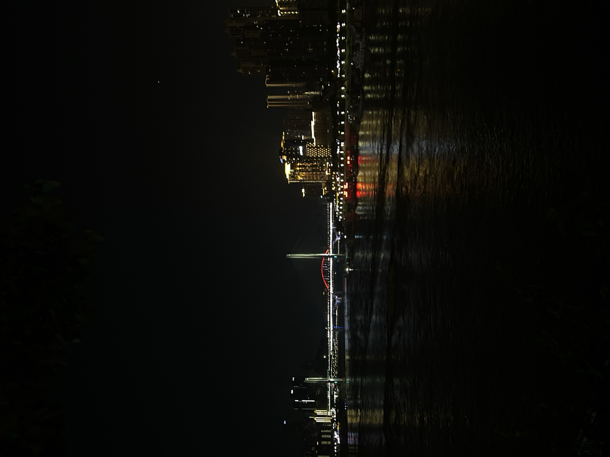 An illuminated cityscape at night, featuring a wide river in the foreground and a bridge spanning across it. The cityscape comprises numerous tall buildings with lights on, indicating a densely populated urban area. The bridge is a prominent structure, also lit up with various colors, including red arches and white supports. Several boats are visible on the river, some appearing to be docked near the shore. The water reflects the city lights, creating a shimmering effect. The sky is dark, typical of nighttime, with a faint star visible. In the foreground, there are dark shapes that appear to be trees or foliage, partially obscuring the view. The image captures a vibrant and active urban environment at night. This view is likely from the Nan'an District, China, looking across the river towards the city.