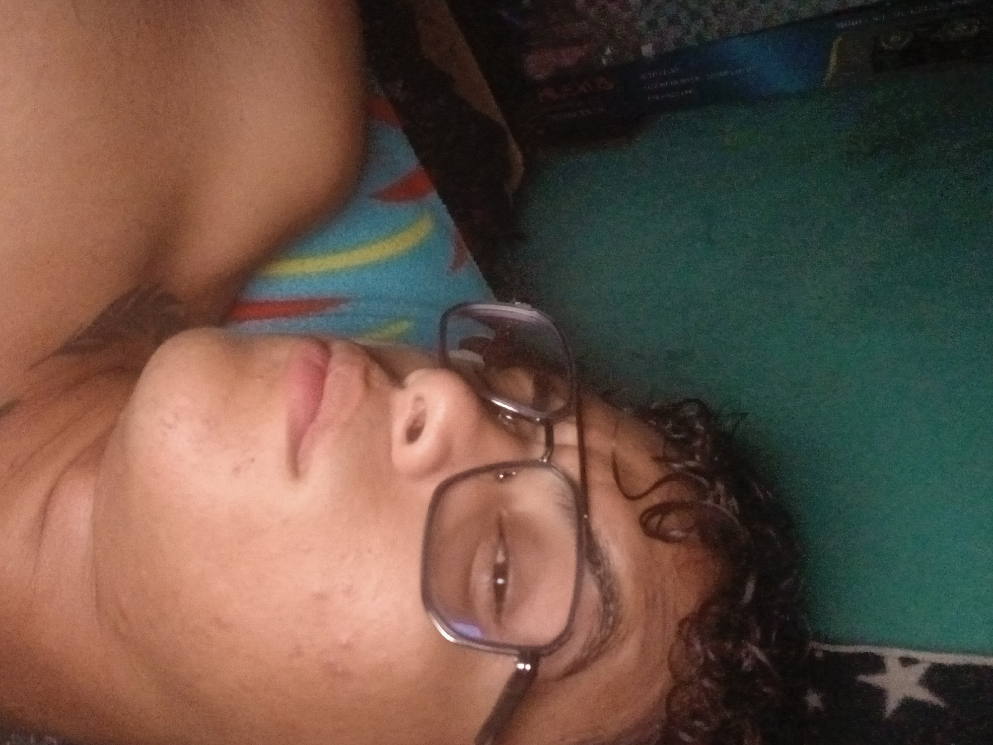 A man is lying down, wearing glasses. He is partially undressed, exposing part of his chest and a tattoo on his shoulder. He appears to be lying on a patterned surface, possibly a bed or couch. The background includes a box with text (partially obscured). The location and city of Altagracia, Venezuela, cannot be determined from the image.
