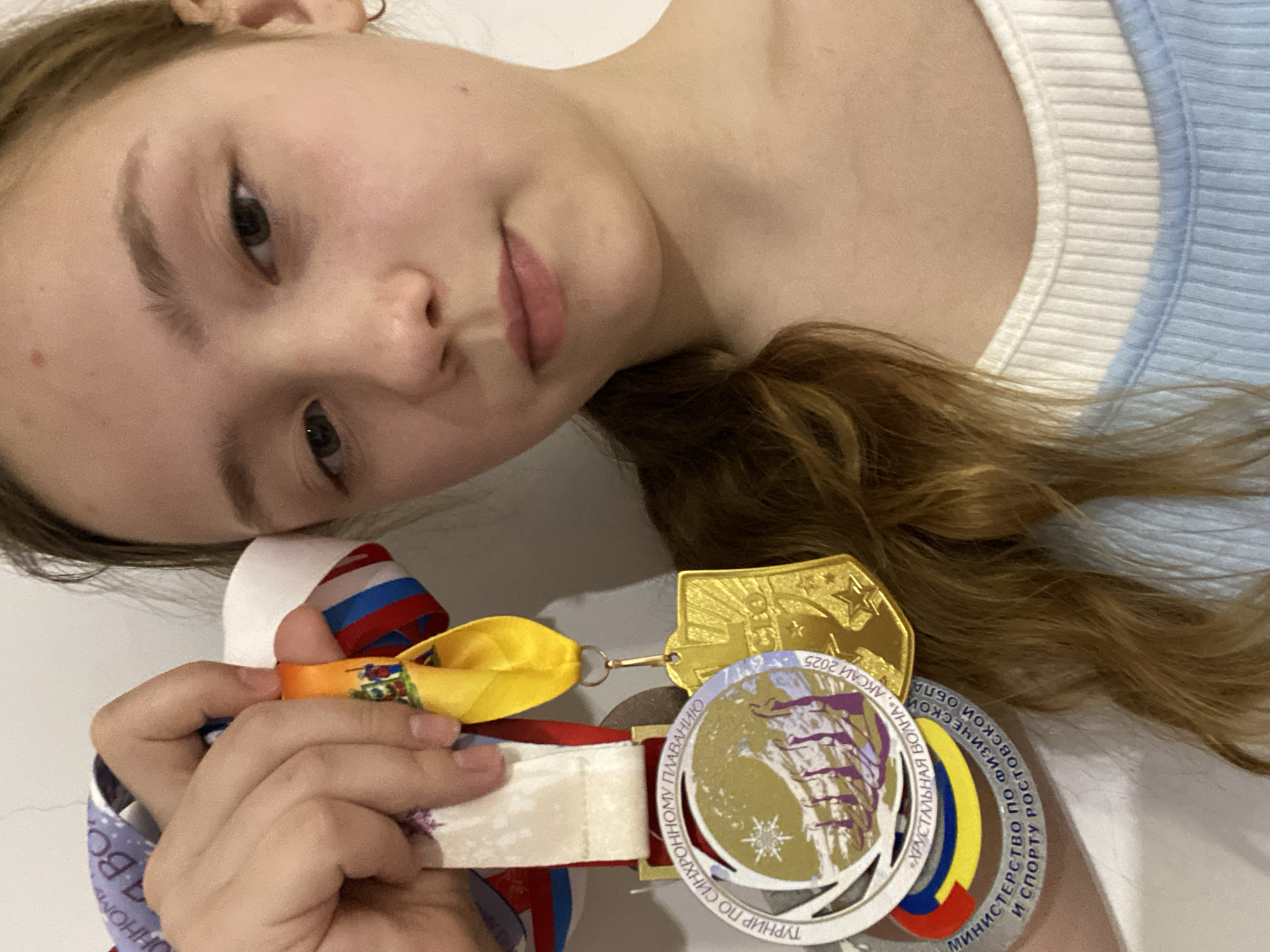 A girl holds a collection of medals. One medal reads "ТУРНИР ПО СИНХРОННОМУ ПЛАВАНИЮ
«ХРУСТАЛЬНАЯ ВОЛНА», АКСАЙ 2025". Another medal reads "МИНИСТЕРСТВО ПО ФИЗИЧЕСКОЙ
И СПОРТУ РОСТОВСКОЙ ОБЛА". The location is Unknown, Unknown.
