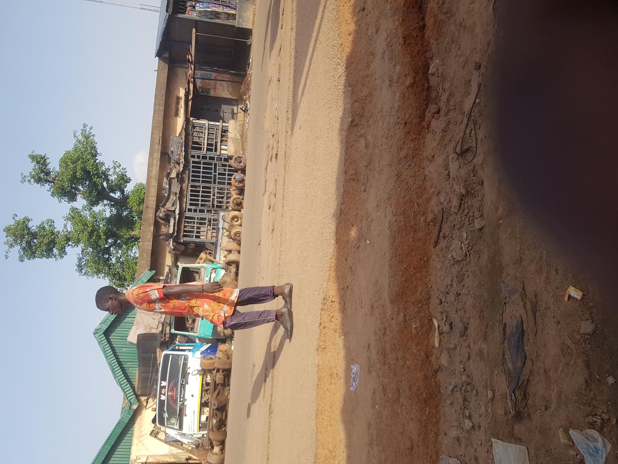 A young man in a patterned orange shirt and dark pants walks along a dirt and asphalt road in Kufena, Nigeria. He is facing to the right and casting a shadow to his left. In the background, there are several disassembled truck chassis and truck parts arranged in front of a low-rise building with a green corrugated roof. The sky is clear and blue. The foreground shows a dirt embankment.