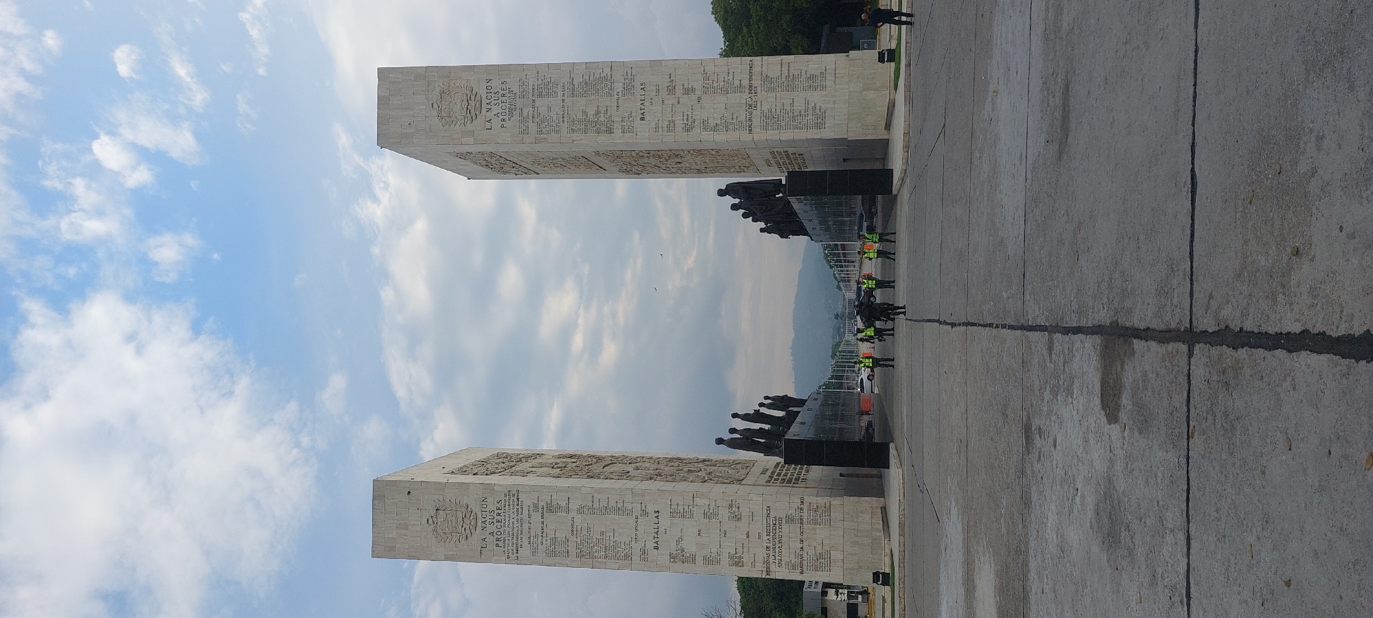 The image captures a monumental structure under a partly cloudy sky, featuring two prominent, tall rectangular obelisks made of light-colored stone, flanking a central, darker base adorned with numerous sculpted figures. The scene is set on a wide concrete plaza, with mountains visible in the hazy distance, characteristic of Caracas, Venezuela.
The obelisks are extensively inscribed with text and carved reliefs. On both obelisks, the phrase "LA NACION A SUS PROCERES" (The Nation to its Heroes) is clearly visible at the top, accompanied by an emblem. Below this, lists of names and categories are etched, including "GENERALES DE BRIGADA" (Brigadier Generals) and "CORONELES" (Colonels), followed by sections titled "BATALLAS" (Battles). One obelisk also lists "HEROINAS DE LA RESISTENCIA Y LA INDEPENDENCIA" (Heroines of the Resistance and Independence) covering "SIGLO XVI, XVII Y XVIII" (16th, 17th, and 18th centuries), and references a specific historical event: "BARINAS 18 DE OCTUBRE DE 1811". The other obelisk similarly mentions "HEROINAS DE LA INDEPENDENCIA".
Between the two obelisks, at a lower level, a dark base features a line of many sculpted human figures, appearing to be standing in formation, possibly representing historical figures or soldiers. On the concrete ground in the foreground, several people are present, including a group of individuals wearing bright green or yellow reflective vests and dark uniforms, possibly security personnel or ceremonial guards, standing in a row facing the monument. Other people are visible in the background near a road or pathway. The time of day appears to be bright daylight, with soft shadows suggesting late morning or early afternoon. The concrete ground shows expansion joints and some marks.