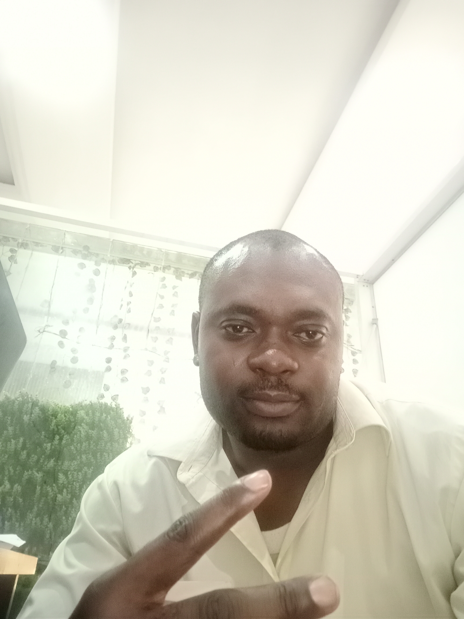 A man is pictured from a low angle. He appears to be in an indoor location with white walls and ceiling panels. He is Black, clean-shaven on top of his head, with a short beard, wearing a light-colored collared shirt. He is making a hand gesture with his right hand. Behind him is a window with a sheer curtain, and a potted plant is partially visible in the background. There is no specific evidence in the photo that indicates it was taken in Gwarinpa, Nigeria, other than an assumption that might be made based on his appearance.