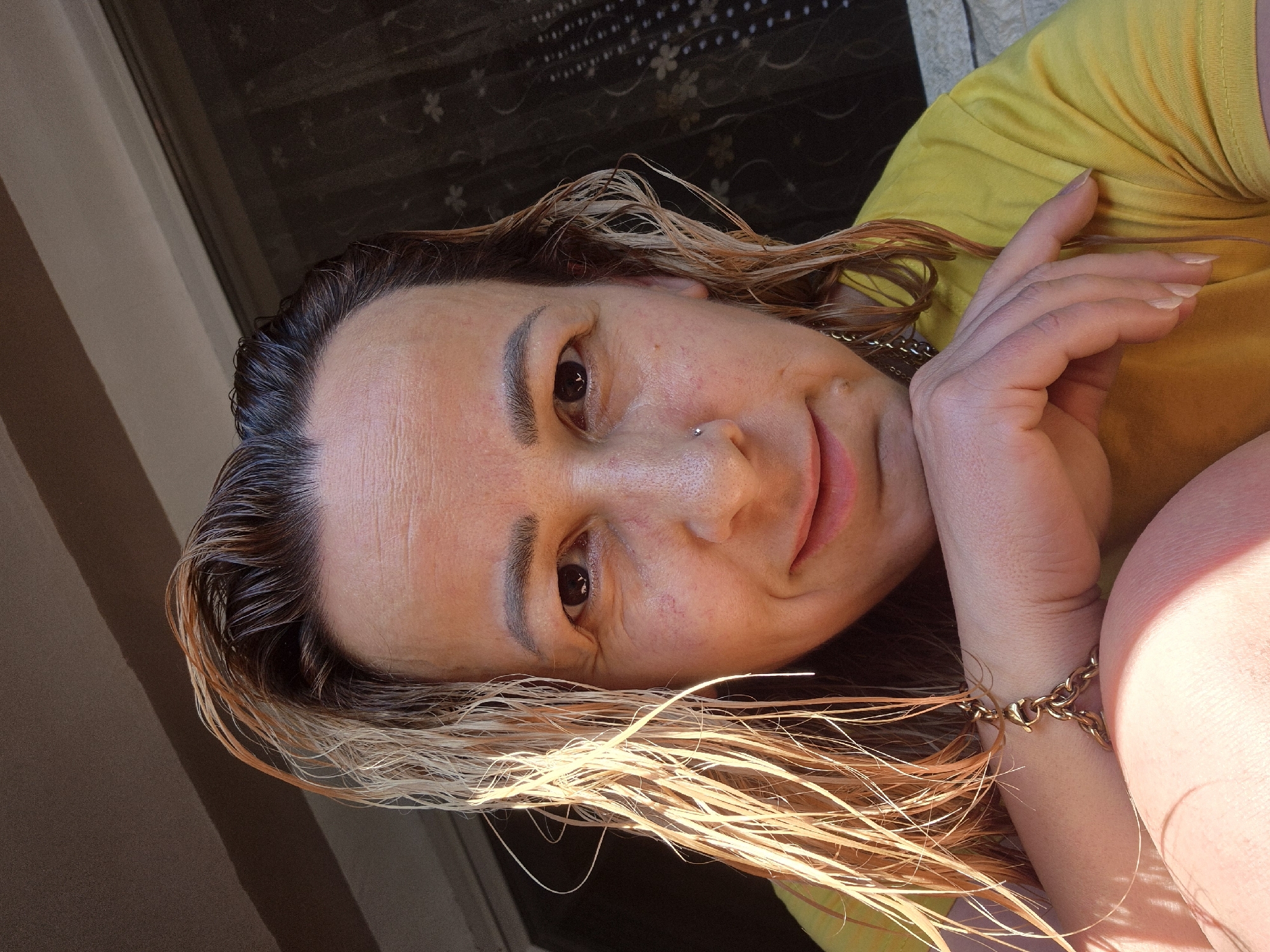 A close-up, front-facing selfie captures a person with wet, blonde-streaked brown hair and fair skin. The individual is wearing a yellow t-shirt and a gold chain bracelet. Their left arm is bent, with the hand resting on their chin, and their right hand is visible supporting their head. A nose ring is visible on the person's left nostril. The background consists of a dark curtain and a window. The lighting suggests an outdoor setting during the daytime. The location is unknown.