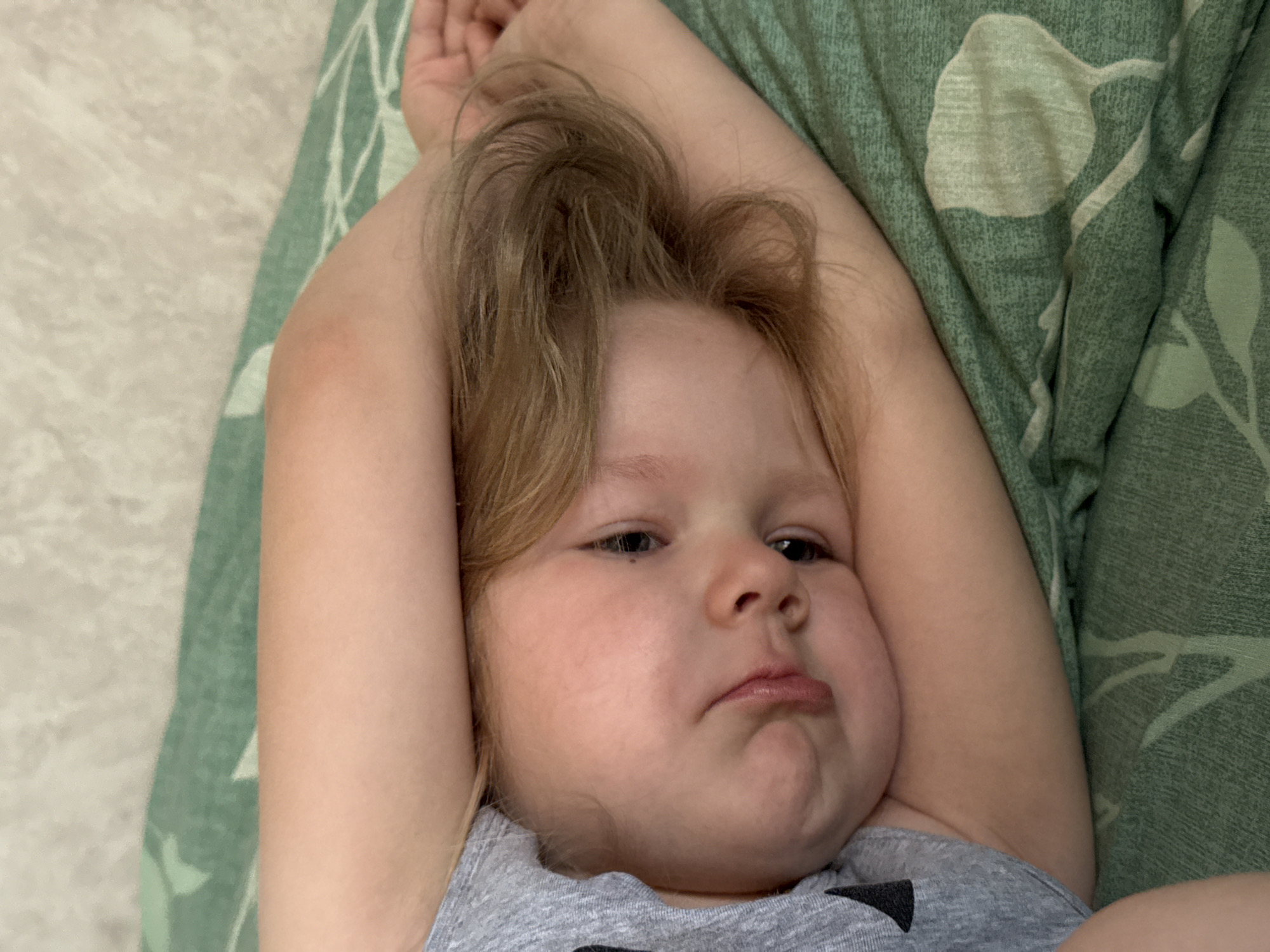 A young child lies on a green patterned fabric, possibly a bed or couch. The child has blonde hair, fair skin, and is wearing a grey shirt. The child's arms are raised, positioned near their head. The facial expression appears sulky or pouting. The location is listed as Ферма, Russia, but there is no element in the image to confirm that.