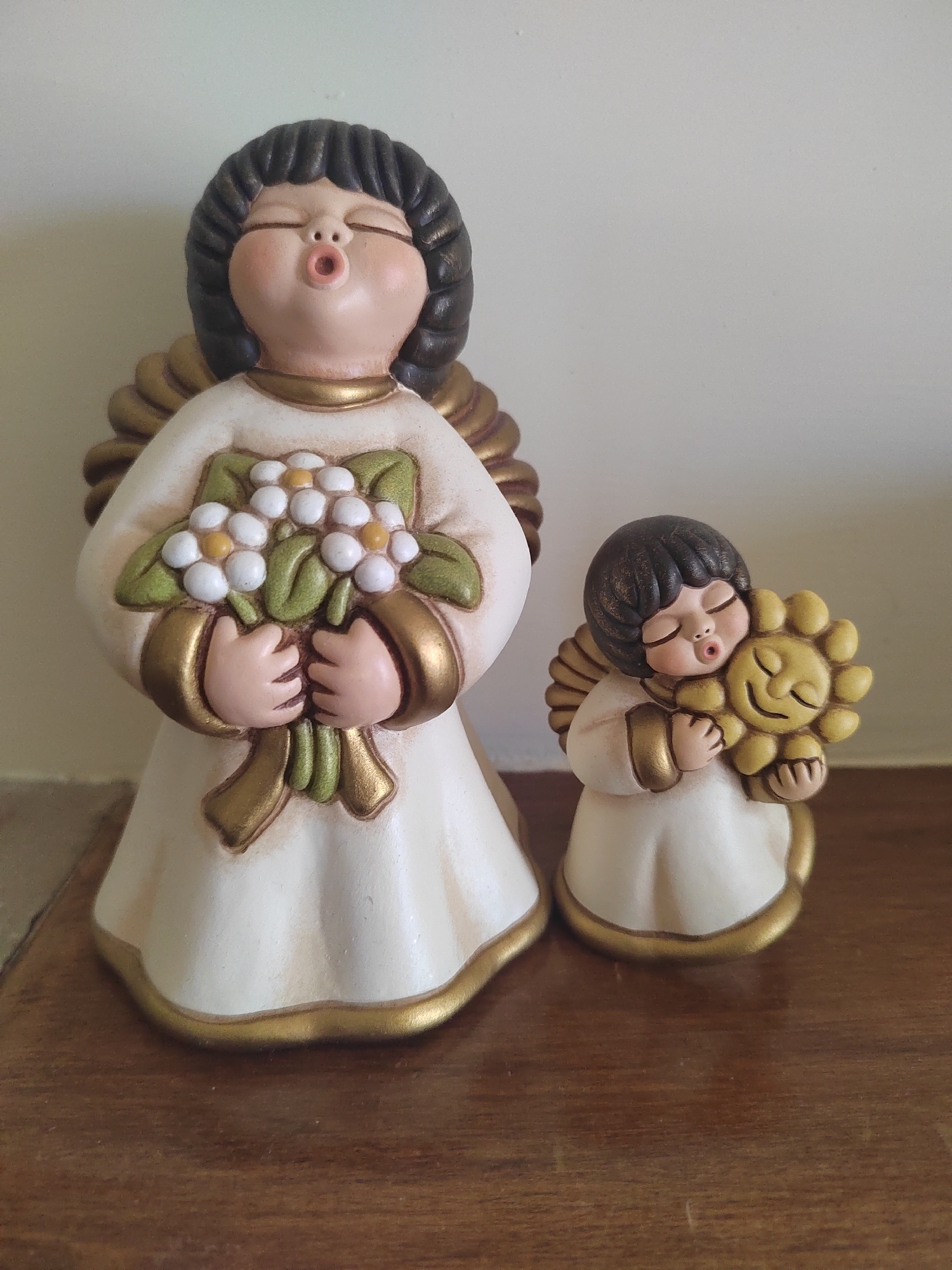 Two ceramic angel figurines are displayed on a wooden surface against a light-colored wall. The larger figurine, on the left, is standing upright with its eyes closed and mouth open as if singing or blowing. It holds a bouquet of white flowers with yellow centers and green leaves. The smaller figurine, on the right, is shorter and is embracing a smiling sun. Both figurines wear white robes trimmed with gold and have dark hair and gold wings. The wooden surface appears to be a table or shelf, showing grain and some subtle imperfections. The background wall is plain and light-colored. There are no individuals present, and the setting does not provide specific geographical markers to pinpoint its location within Porto Recanati, Italy.