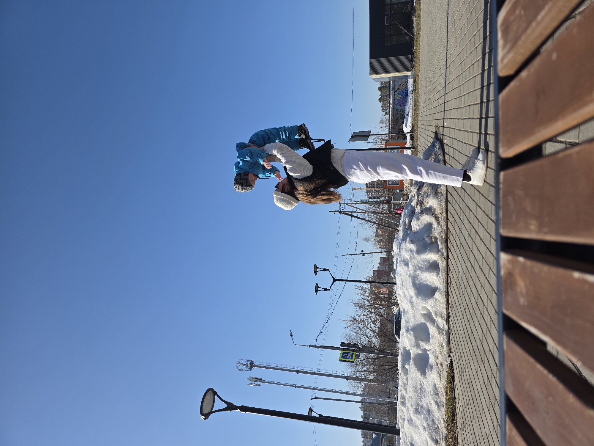 A person in white pants, white shoes, a white sweater, and a black vest is holding a child in a blue snowsuit and gray knit cap upside down in the air. The background features a clear blue sky, a paved walkway, a wooden bench, snow-covered ground, leafless trees, streetlights, power poles, buildings, and a road with a pedestrian crossing sign. The location appears to be Chaikovsky, Russia.