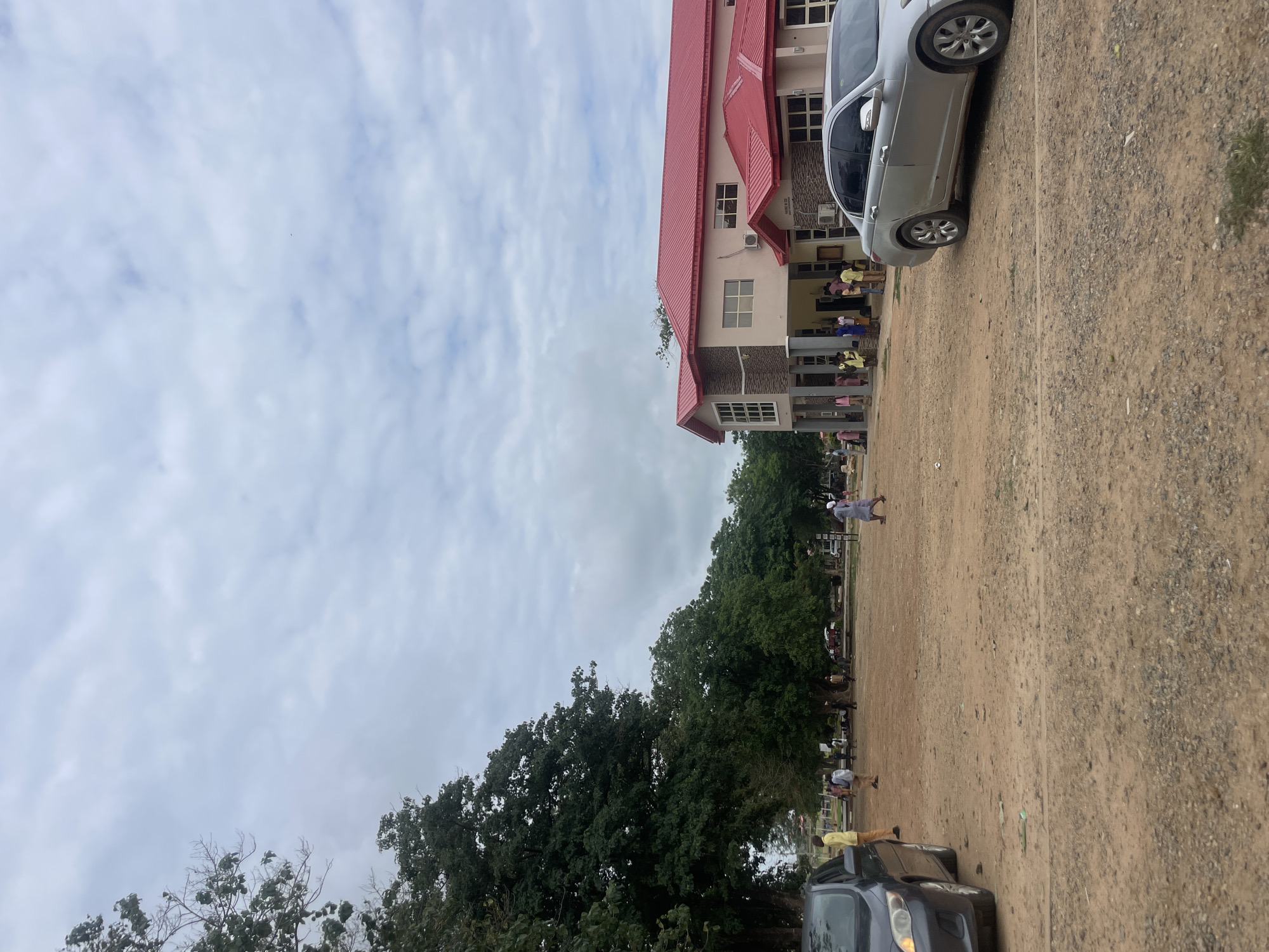 This image depicts an outdoor scene in Karu, Nigeria, characterized by an unpaved, light brown dirt and gravel ground surface with scattered small patches of green vegetation. The sky is overcast with light grey clouds.
On the right side of the frame, a two-story building with a prominent bright red corrugated metal roof is visible. The building's walls are light beige, with sections of grey stone cladding on the lower level and around window frames. Multiple rectangular windows are present, and a front entrance area features structural columns and an overhang. Two external air conditioning units are mounted on the building's facade.
To the left, a dense line of large, leafy green trees extends from the midground into the background, with some bare branches visible in the upper left corner.
Two sedans are present: a dark grey sedan is partially visible on the far left, facing towards the right. A silver sedan is parked on the right side, facing towards the left, with its side profile prominent.
Approximately 15-20 individuals are dispersed across the scene. A group of people, including adults and several younger individuals, stands near the front entrance of the building on the right. Other individuals are seen walking across the open dirt area; for example, one person in a yellow top and dark trousers stands near the dark grey car on the left, and another person in a light grey tunic and white head covering walks towards the left on the right side of the frame. Clothing visible includes tunics, trousers, and dresses in various colors.