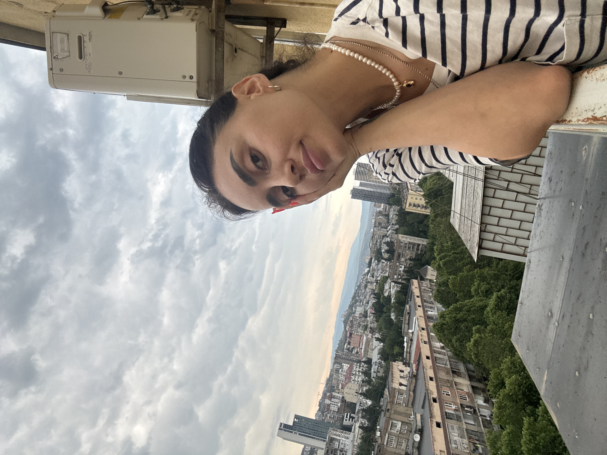 In an outdoor, slightly overcast urban scene in Tbilisi, Georgia, a young woman is captured in a selfie-style shot from the chest up. She is positioned on the right side of the frame, leaning on a railing with her right arm and hand supporting her face. Her left arm is bent at the elbow and rests on the railing. She has dark hair pulled back and is wearing a striped t-shirt and a pearl necklace with a pendant. In the background, a cityscape unfolds, featuring a mix of apartment buildings, some with balconies, and a taller modern skyscraper on the left. Lush green trees are visible between the buildings, and a hilly landscape with distant mountains can be seen further back under a cloudy sky. An outdoor air conditioning unit is mounted on the wall to the right of the woman.