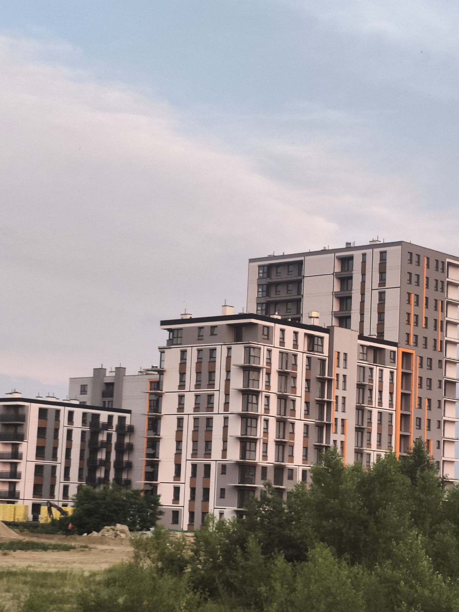 This is a photograph of a modern apartment complex in Nowy Dwór Mazowiecki, Poland. The buildings are tall and multi-story, featuring a geometric design with alternating white, grey, and brown facades. Many windows and balconies are visible, some with dark frames and others with lighter ones. Vertical orange accents add a pop of color to one of the buildings. In the foreground, lush green trees and vegetation partially obscure the lower levels of the structures. The sky above is a pale blue with scattered clouds, suggesting it is daytime. Some construction materials, including yellow tarps and a pile of dirt, are visible on the ground, indicating ongoing development in the area.