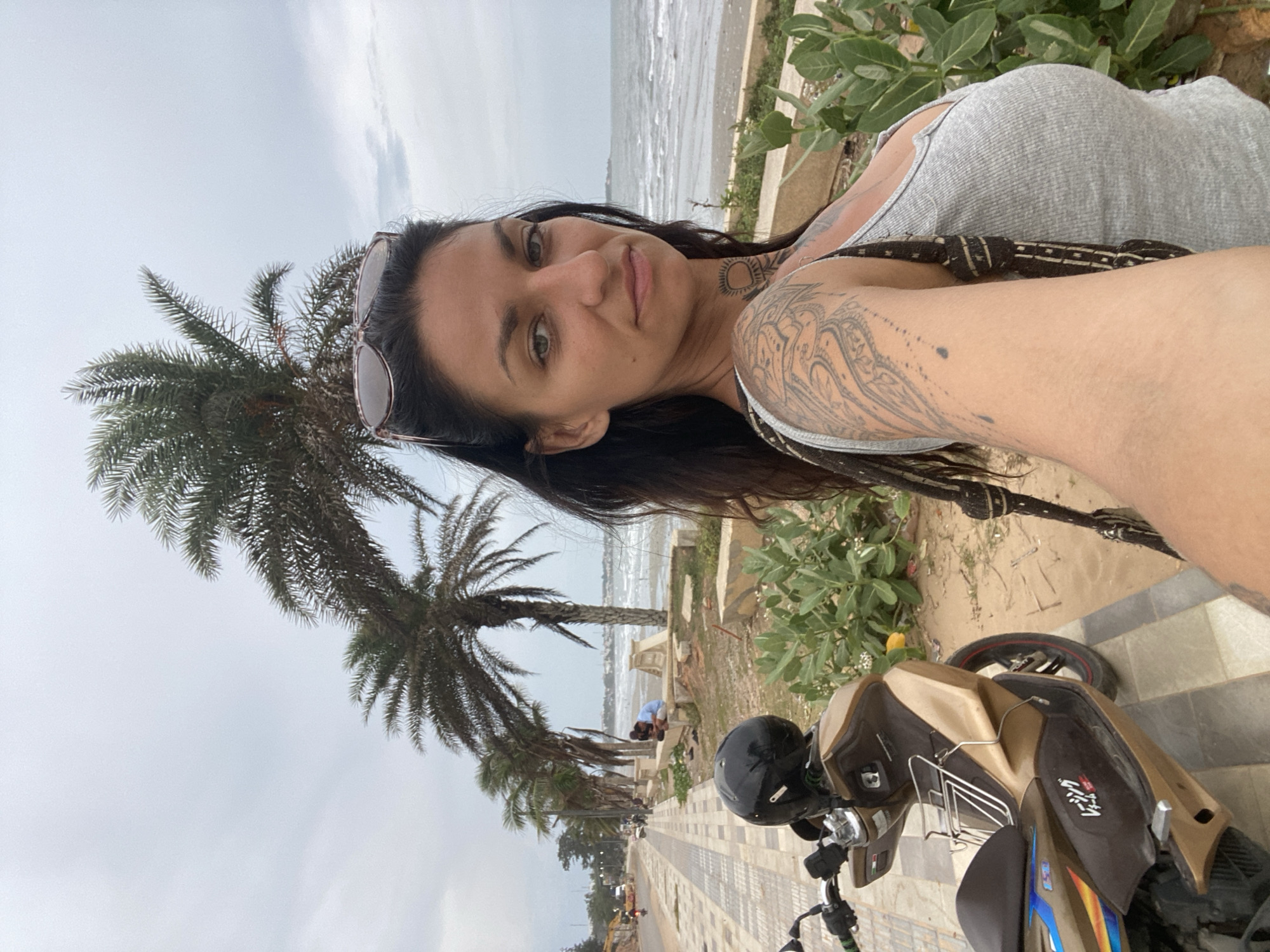 In Mũi Né, Vietnam, a woman with dark hair and extensive arm and neck tattoos takes a selfie on a cloudy day. She wears a grey tank top and sunglasses perched on her head. To her left, a beige and brown scooter with a black helmet rests on a paved walkway. The walkway is lined with palm trees and a low wall bordering a sandy area with green plants. Beyond the sandy area, the ocean stretches to the horizon under an overcast sky. In the background, people are visible sitting on the wall near the water, and a street with a bus and motorcycles is to the left of the walkway.