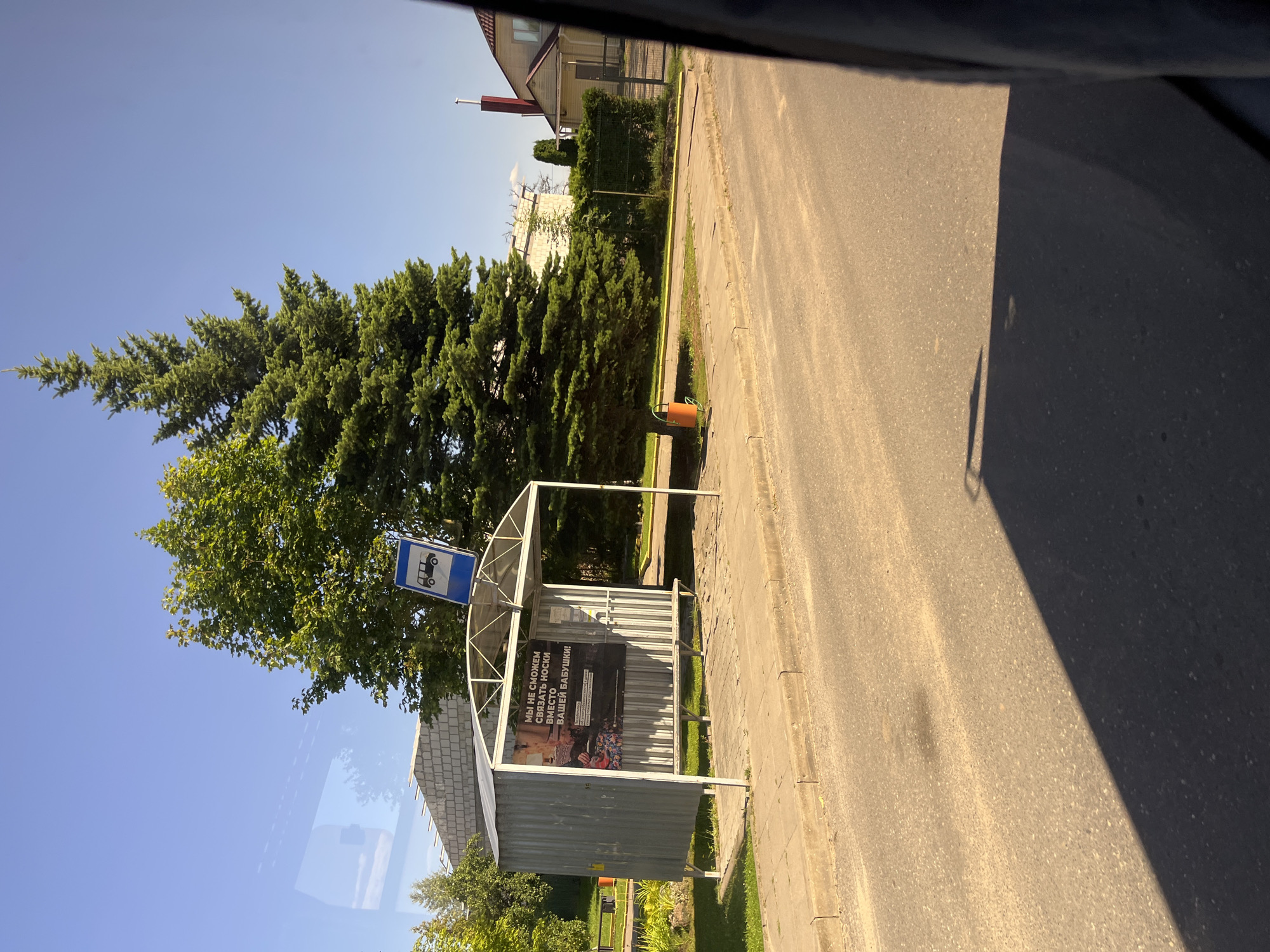 The image depicts a bus stop in Pastavy, Belarus, on a clear, sunny day. The bus stop shelter is constructed of corrugated metal and has a white metal frame. A sign with a blue background and a white bus icon is affixed to the top of the shelter. Inside the shelter, a poster is displayed, featuring Russian text that reads "Мы не сможем связать носки вместо вашей бабушки!" which translates to "We can't knit socks instead of your grandmother!". Below this, there is an image of various knitted items. A wooden bench is present within the shelter.
To the left of the bus stop, a light-colored brick building under construction is visible, with scaffolding. Further left, a lush green tree with dense foliage stands, and beyond that, a smaller evergreen tree. To the right of the bus stop, a tall, mature evergreen tree dominates the scene. In the background, a residential house with a red roof and white siding is partially visible, along with a tall red chimney. A hedge and a metal fence separate the property from the street.
The foreground shows a paved road with a slightly uneven surface and a distinct shadow cast across it, indicating the sun's position. A concrete sidewalk runs along the edge of the road, next to the bus stop. A bright orange trash bin is situated near the base of the large evergreen tree. The sky is a clear, vibrant blue.