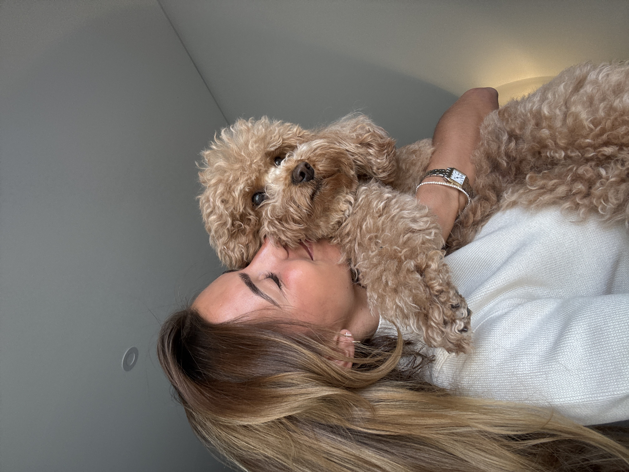 A woman lies on a white textured surface, presumably a bed or sofa, with a tan poodle resting on her chest and neck. The woman has long, light-brown hair with blonde highlights. She wears a white sweater and two bracelets and a watch on her left wrist. Her eyes are closed, and her head is slightly tilted to the side. The dog's face is close to hers, near her mouth, looking directly at the camera. The background is a plain gray wall with a circular light fixture visible in the upper left corner. The location is not discernible as being in Vila Nova de Gaia, Portugal; the environment is generic.