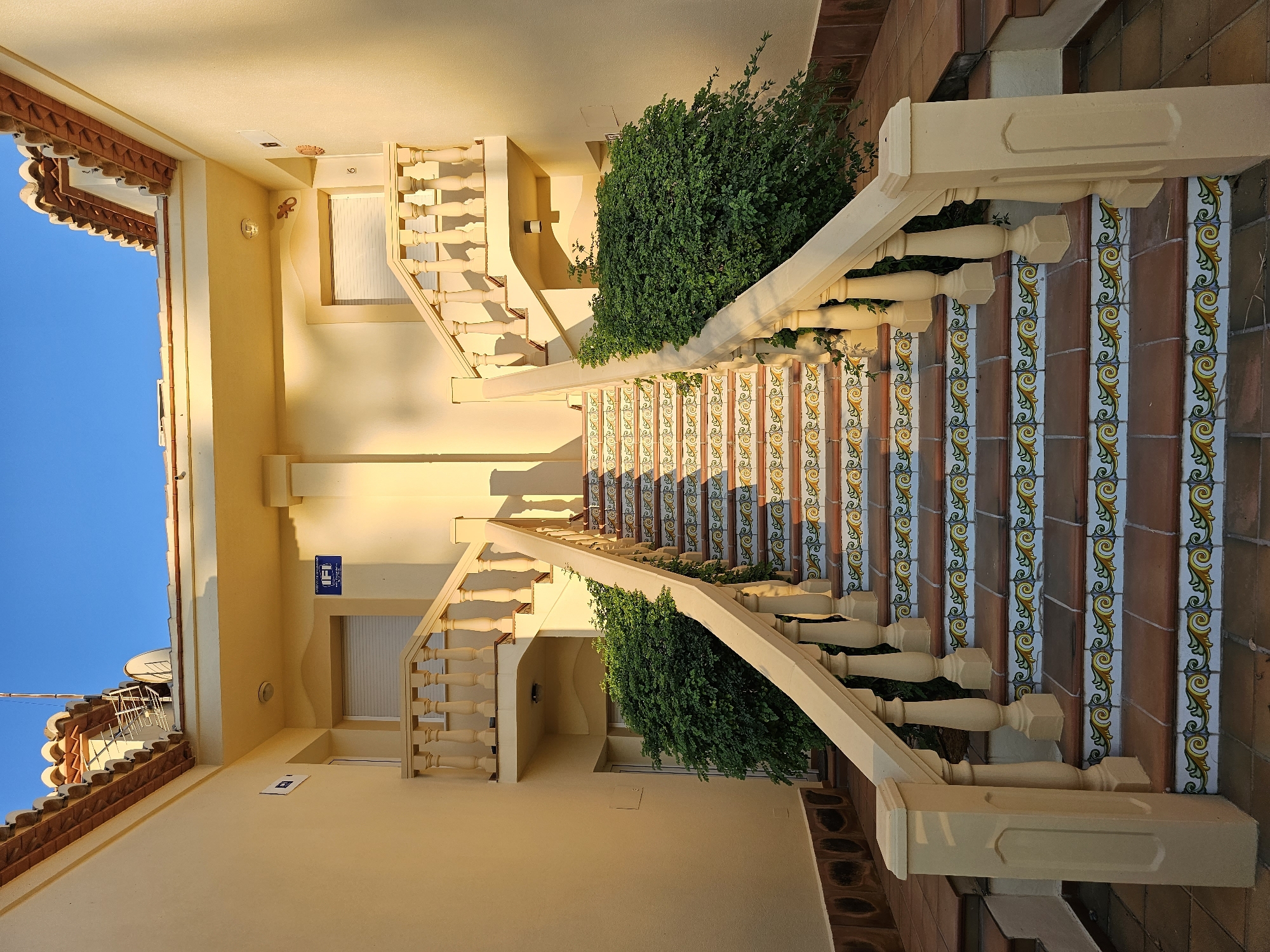 The image shows an external staircase leading to two identical doorways. The staircase has a cream-colored balustrade and is decorated with colorful tiles featuring a yellow, green, and blue ornate pattern. Lush green bushes are planted on either side of the staircase. The building walls are painted a light yellow, and the roof has terracotta tiles. A blue sign with "IP" in white is visible on the left side of the building. The scene is illuminated by sunlight, casting shadows on the stairs and walls. This is likely a residential building or apartment complex in Deveses, Spain.